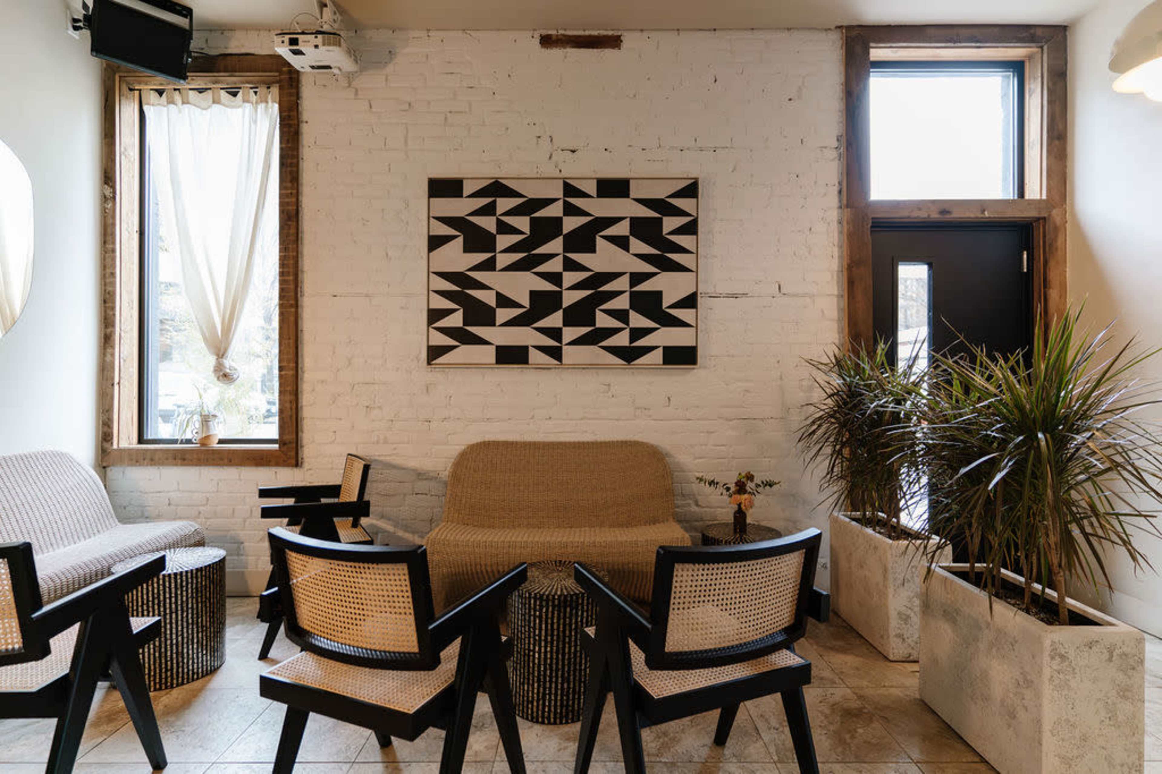 The image shows a cozy interior space with a minimalist design, featuring a large black-and-white geometric artwork on the wall, comfortable seating arrangements, and potted plants.