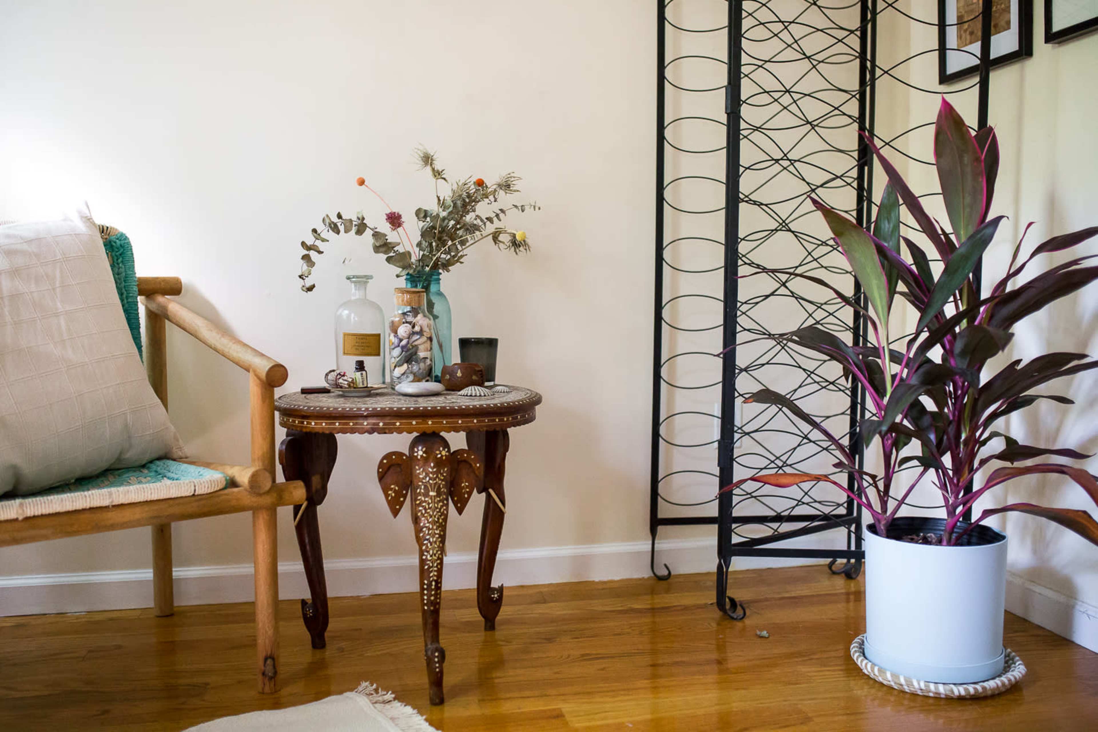 A wooden chair sits beside a round decorative table adorned with a vase and plant, while a metal shelving unit stands against the wall.