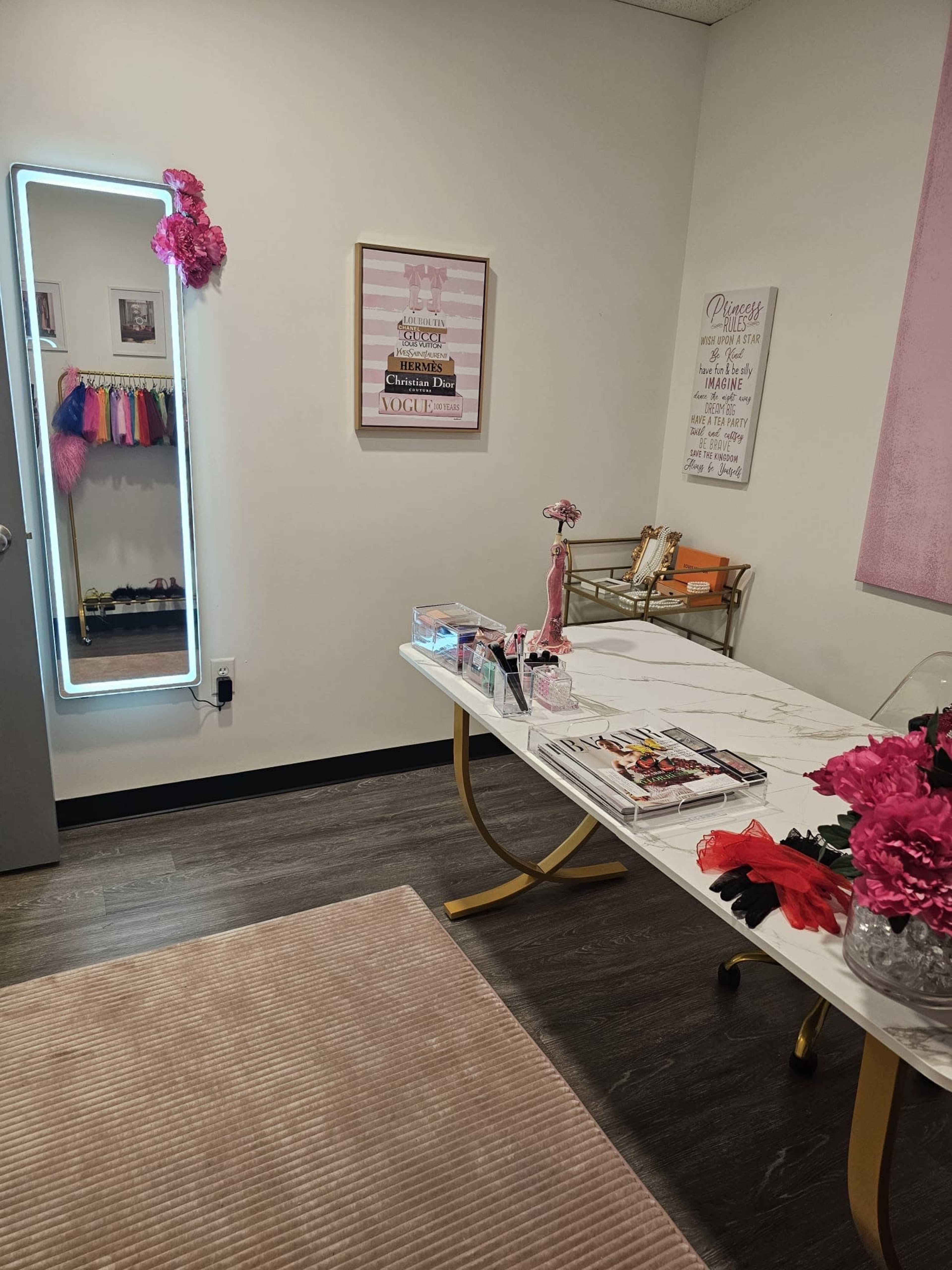 A room with a marble-top table, a large illuminated mirror, and pink decor elements, including flowers and framed artwork on the walls.