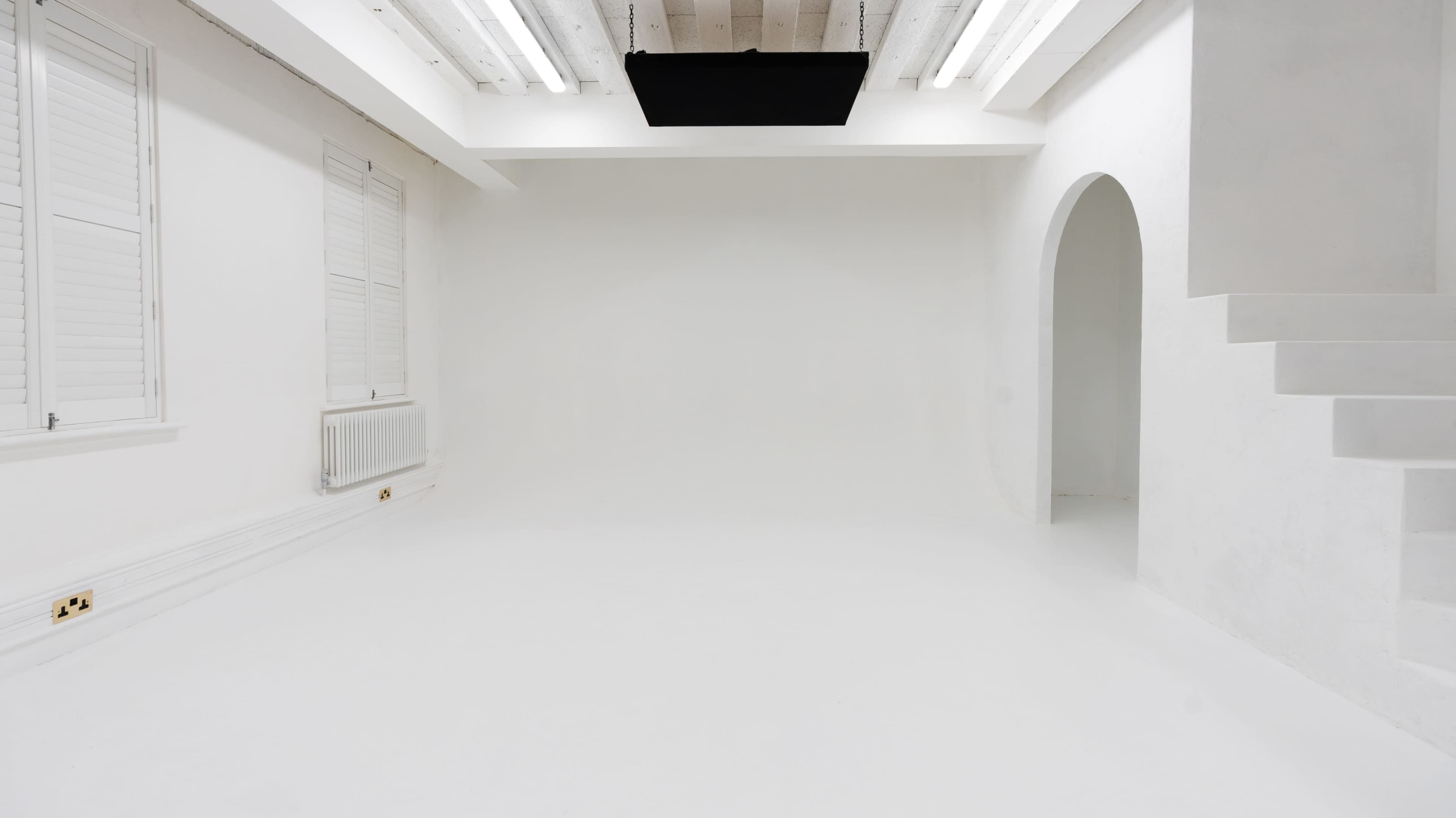 The image features a minimalist white interior with bare walls, a curved archway, and a staircase in one corner.