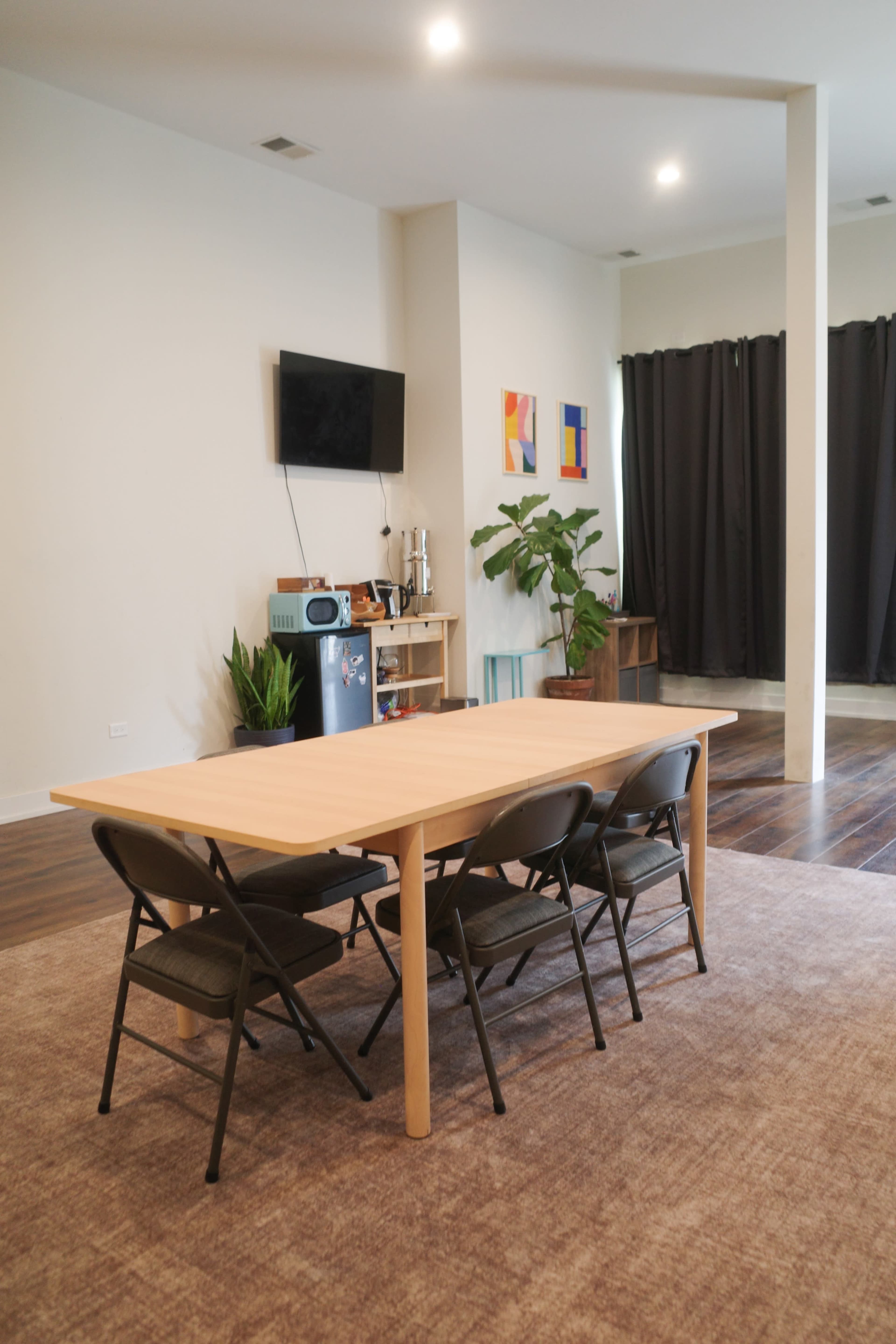 A spacious room features a wooden dining table surrounded by four folding chairs, a television mounted on the wall, and potted plants near a sideboard.