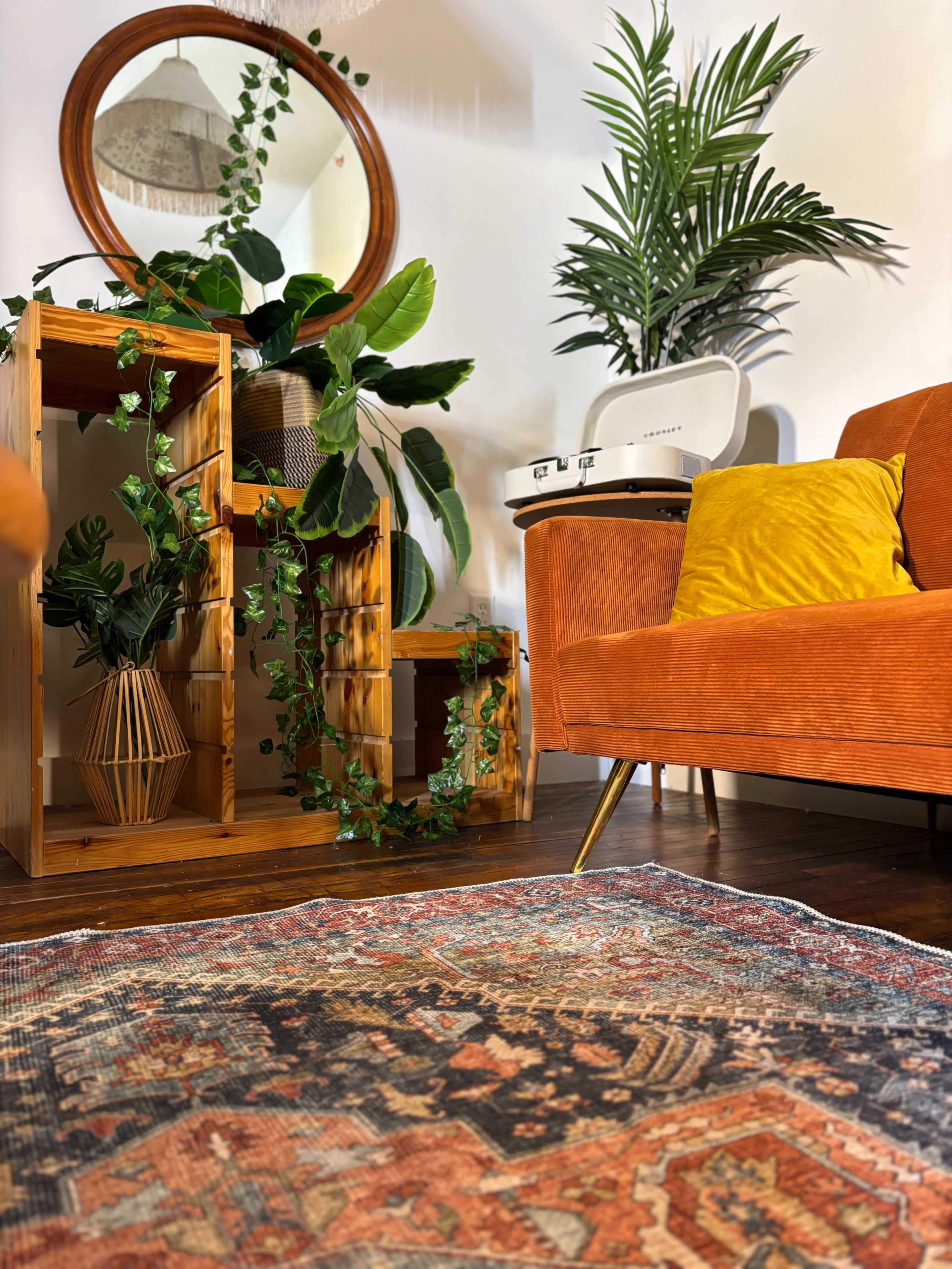 The image features a cozy living room with an orange sofa, a decorative rug, wooden shelves with plants, and a round mirror on the wall.