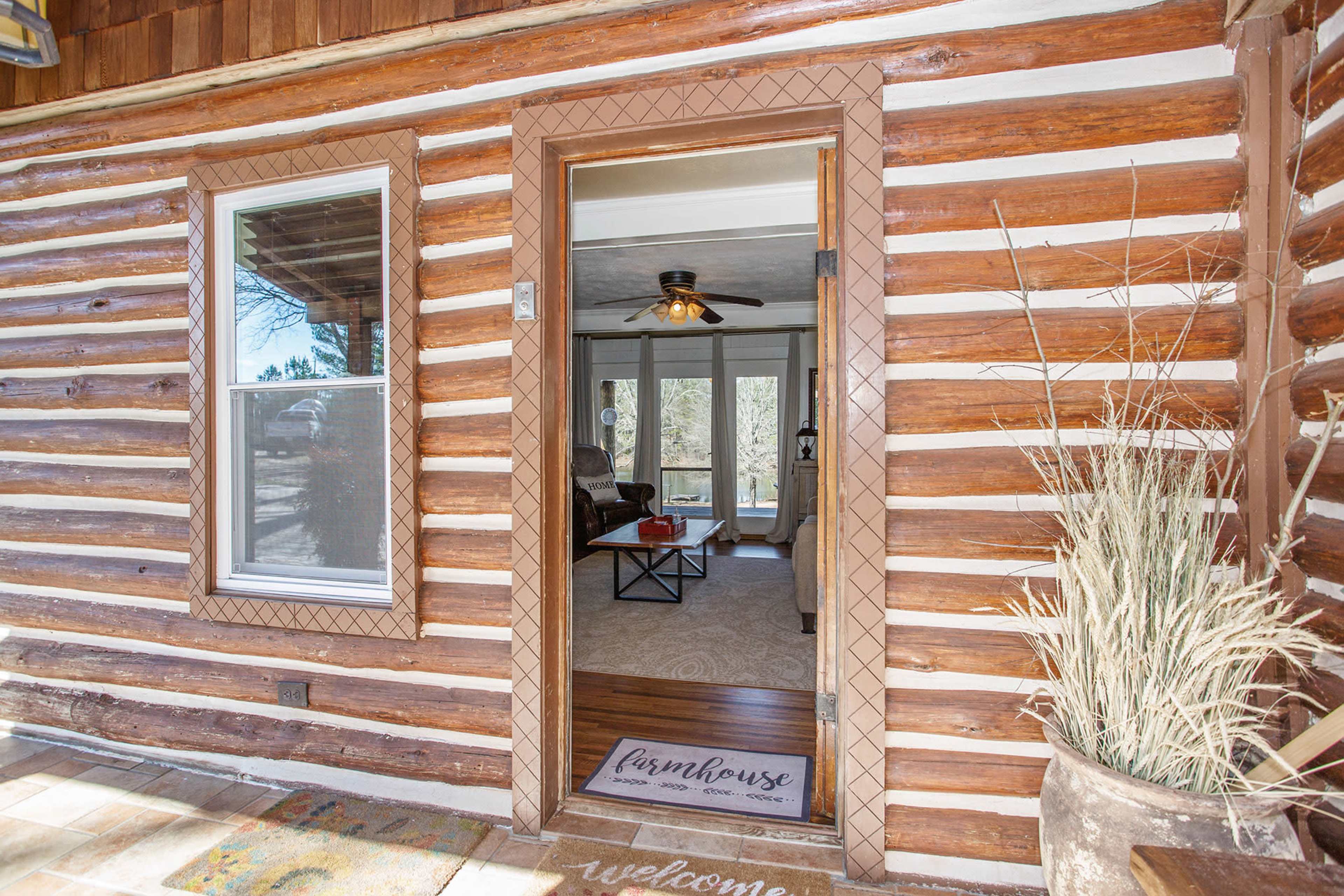 The entrance of a log cabin features a welcome mat and a view into a cozy living area with large windows.
