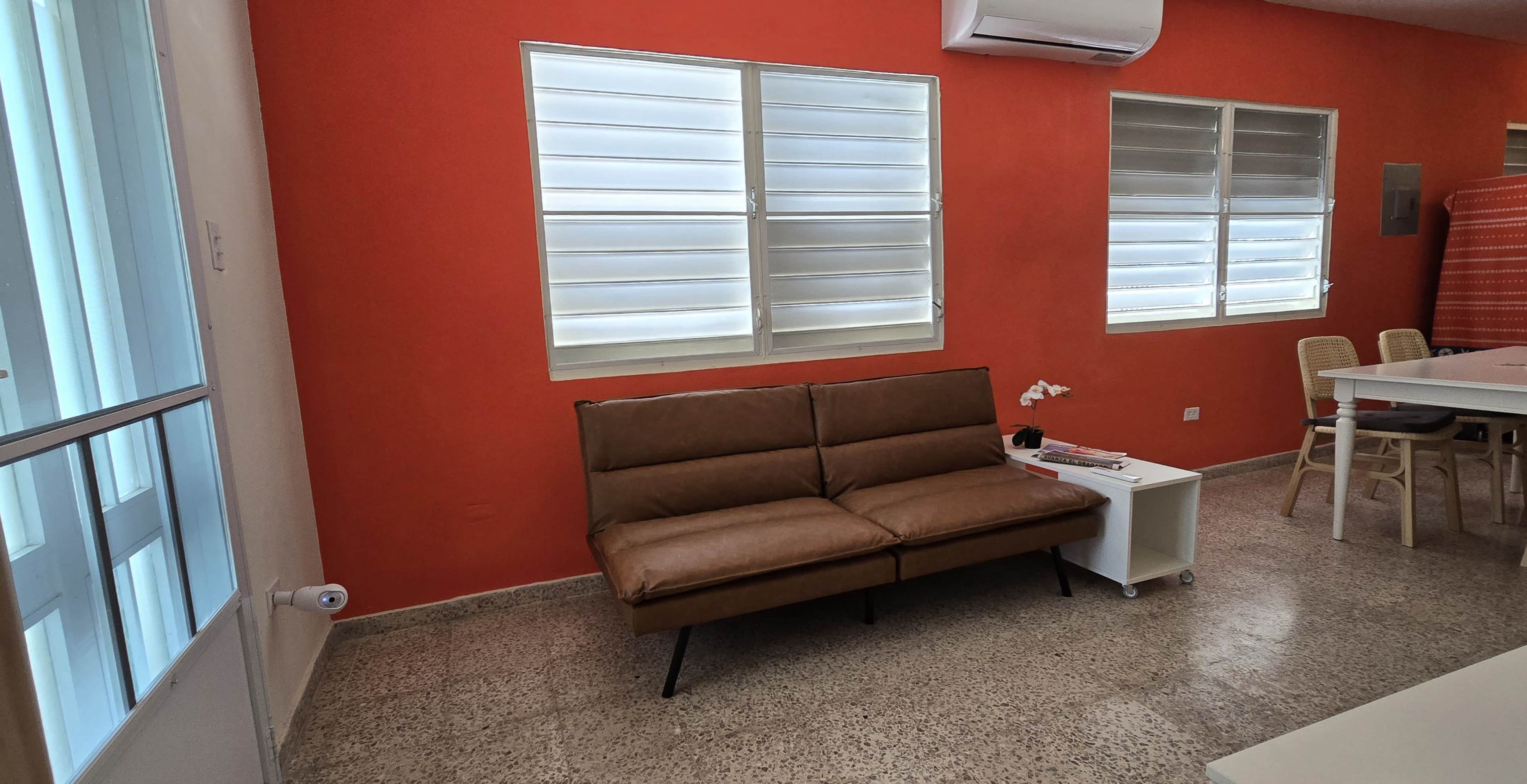 The image shows a room with a red wall, a brown sofa, two windows, and a dining area with white chairs and a table.