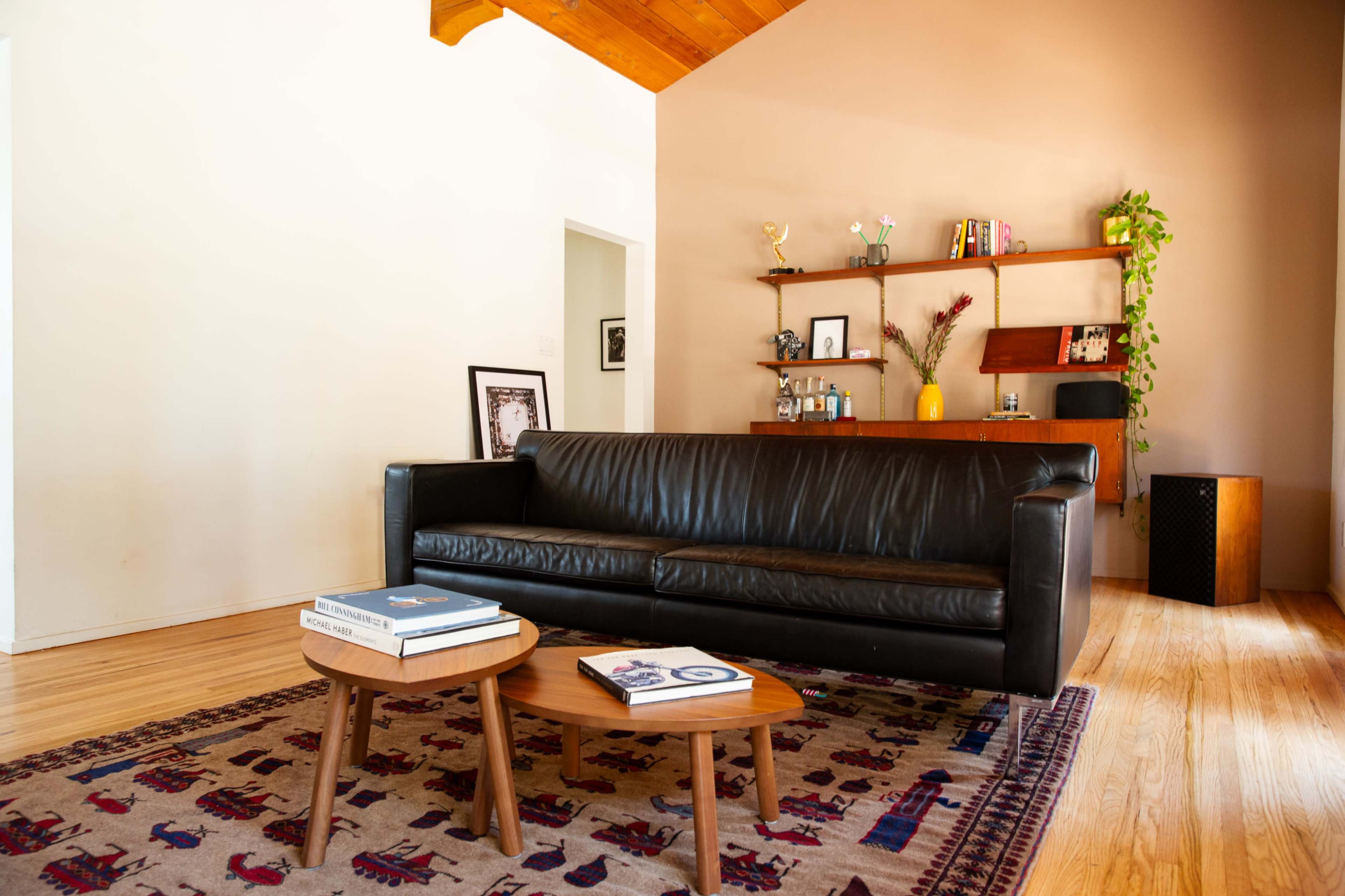 A living room with a black leather sofa, two wooden tables, and a bookshelf featuring decorative items and plants.