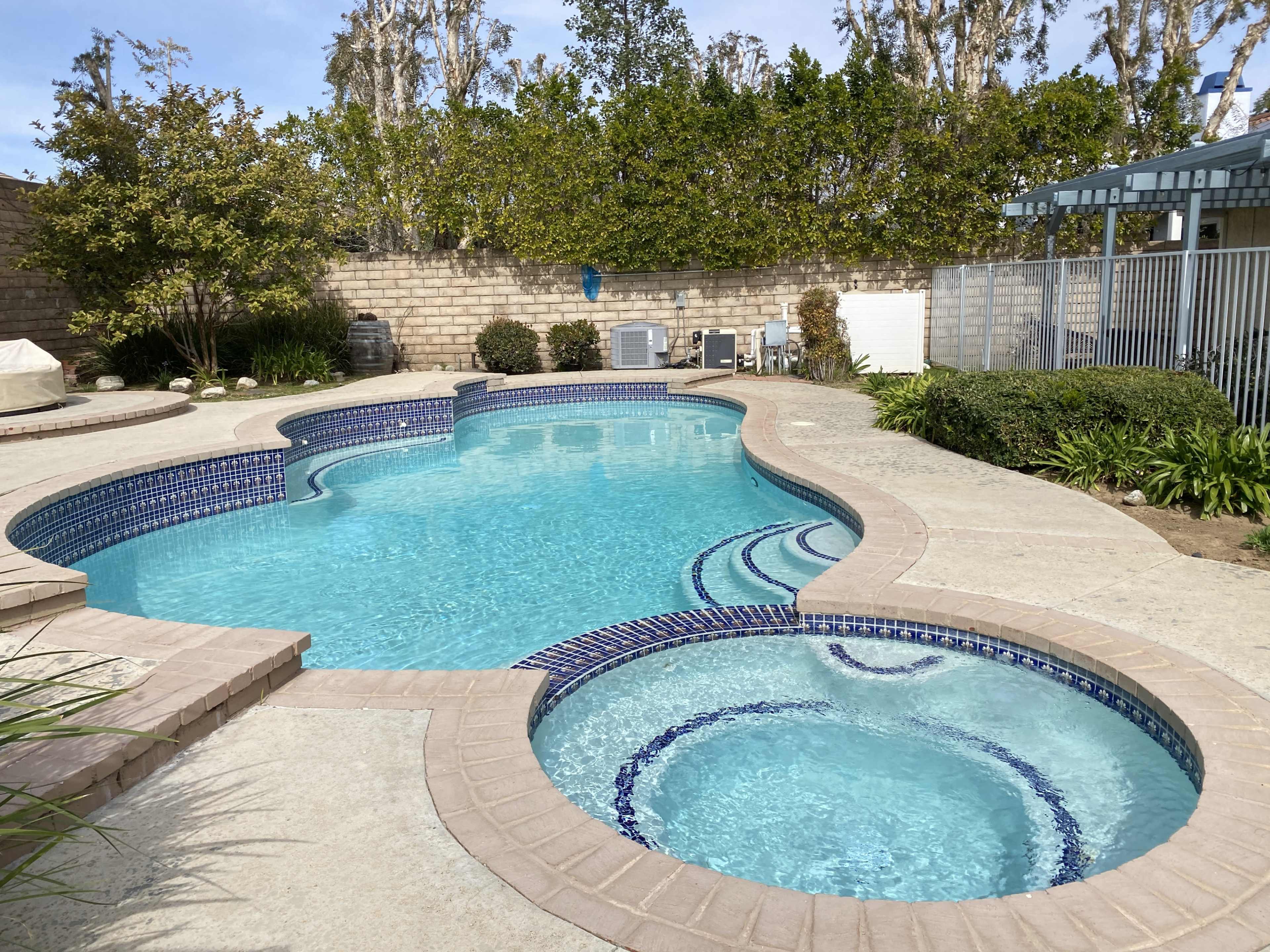 A residential backyard with a swimming pool that features a spa area, surrounded by greenery and lounge chairs.