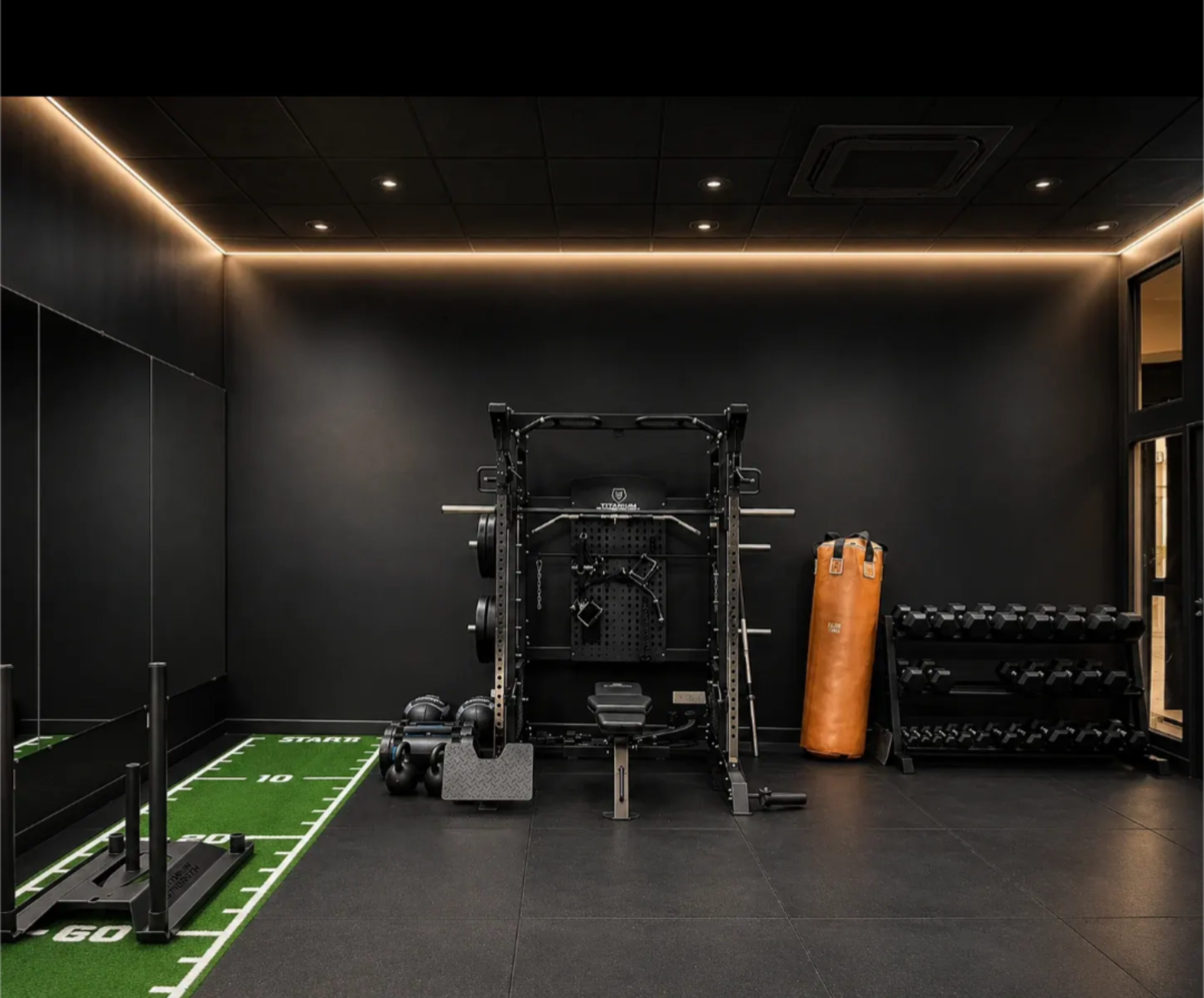 The image shows a modern gym interior featuring a weightlifting station, dumbbells, a punching bag, and a turf area marked with yard lines.