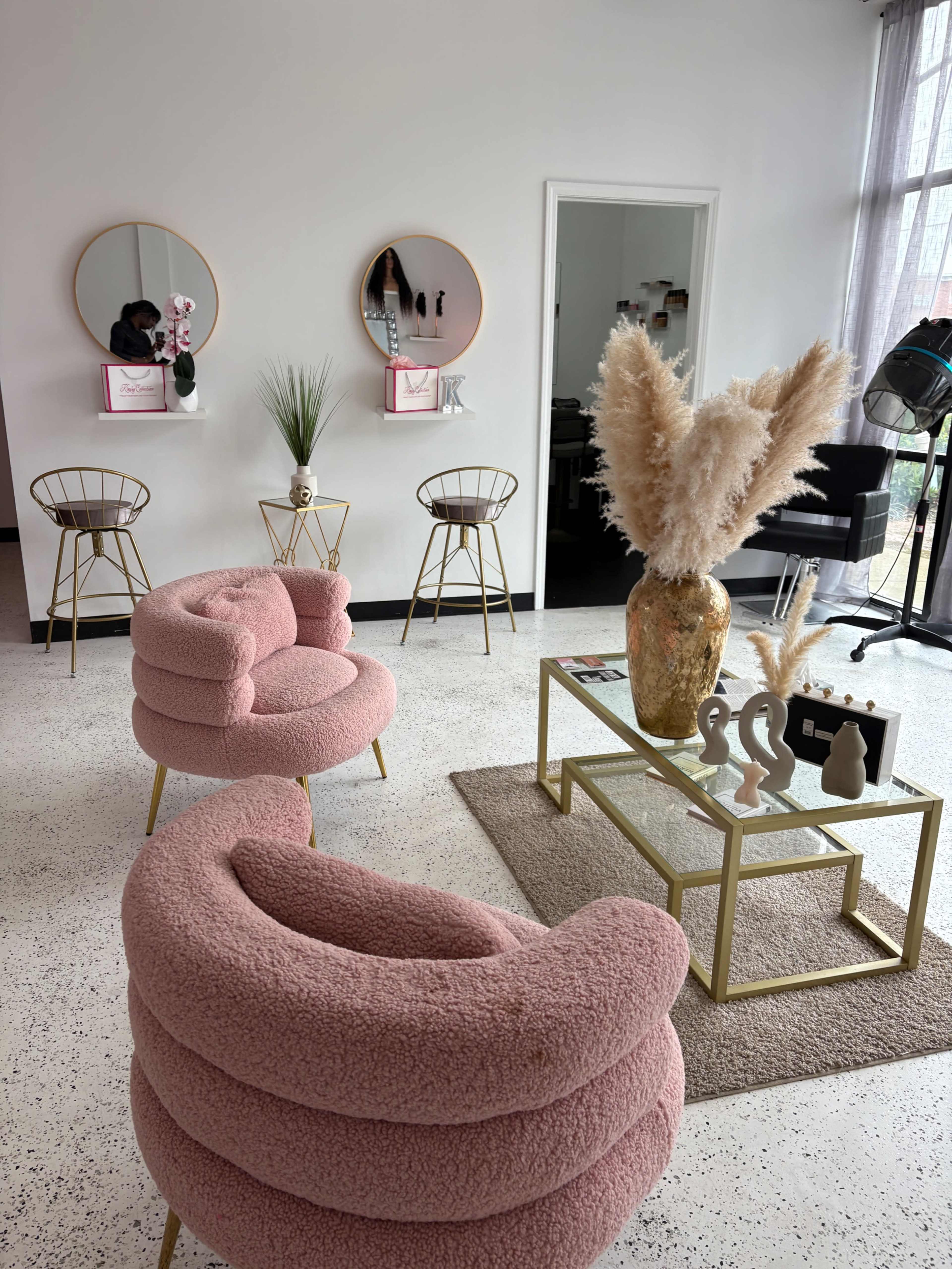 The image shows a modern interior space with pink, textured chairs, gold-accented furniture, mirrors on the wall, and decorative elements, including a tall vase with pampas grass.
