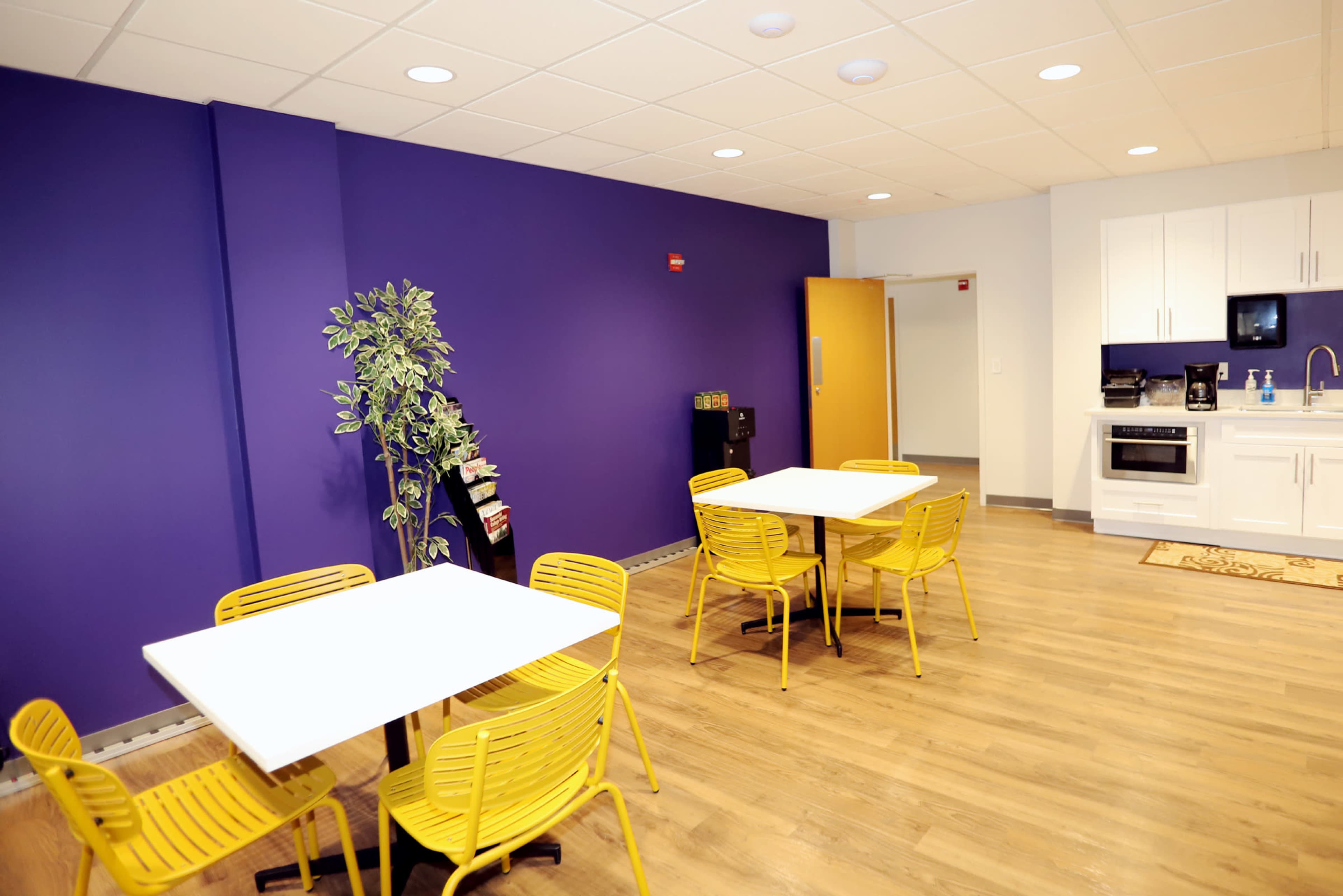 The image shows a modern break room featuring yellow chairs and tables against a purple wall, with a kitchenette area in the background.