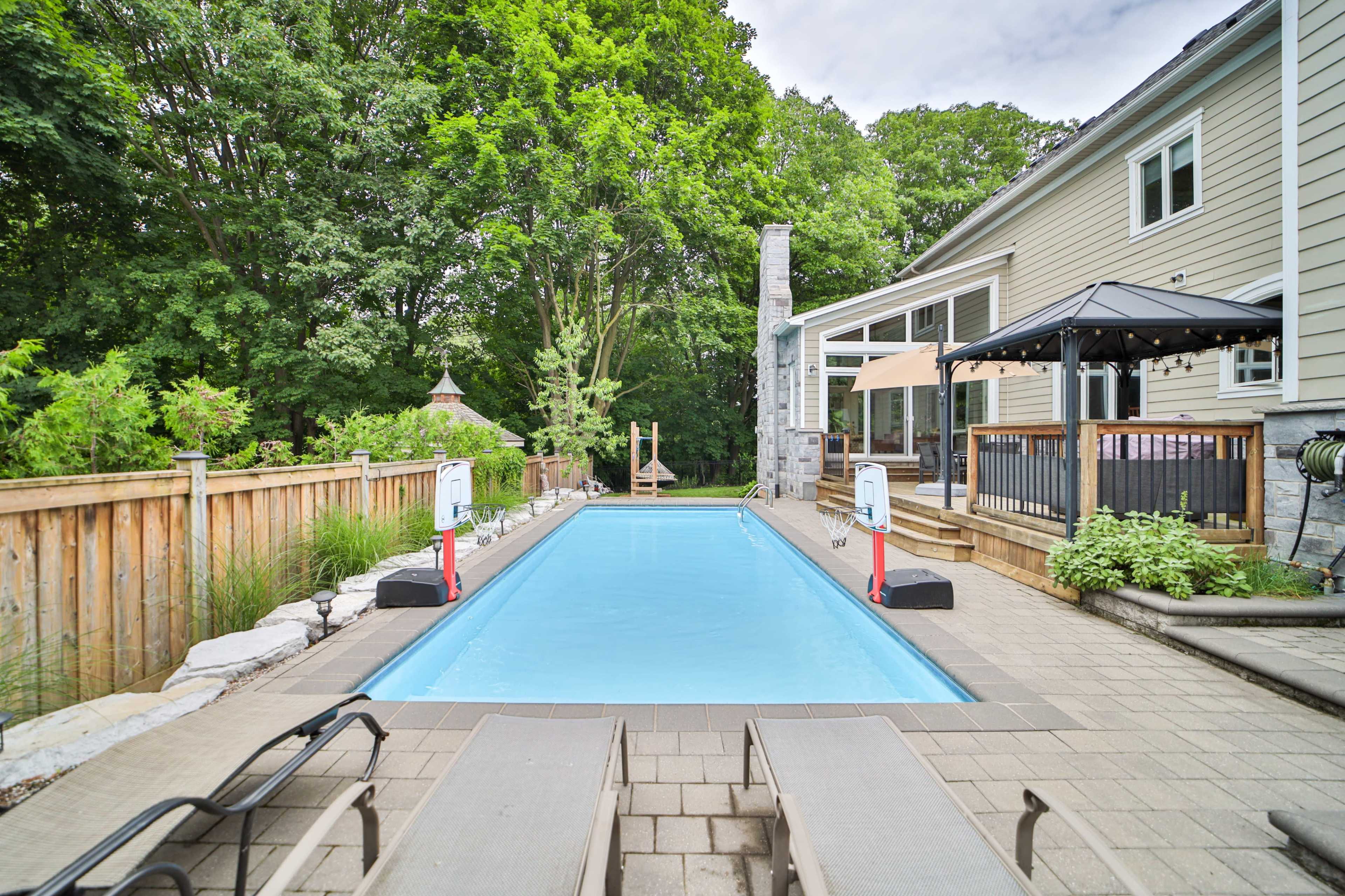 The image shows a rectangular swimming pool surrounded by a stone patio, lounge chairs, and a gazebo, with greenery in the background.
