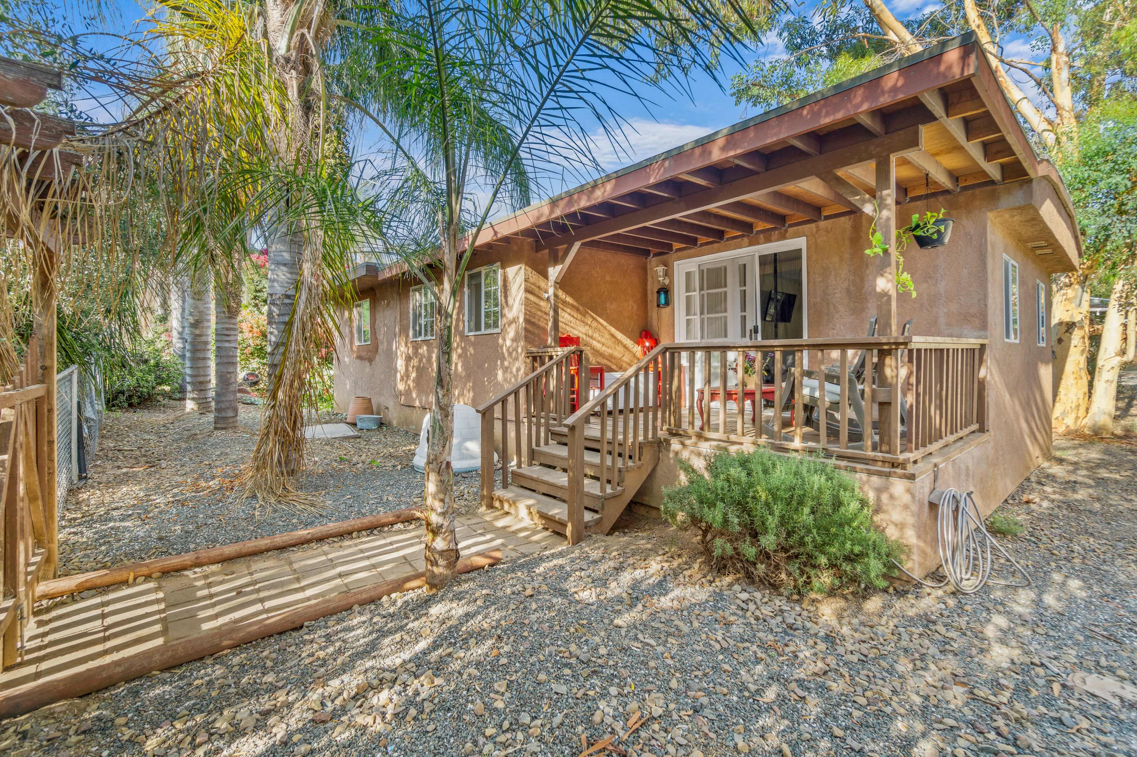 A small, single-story house with a wooden porch is surrounded by palm trees and gravel landscaping.