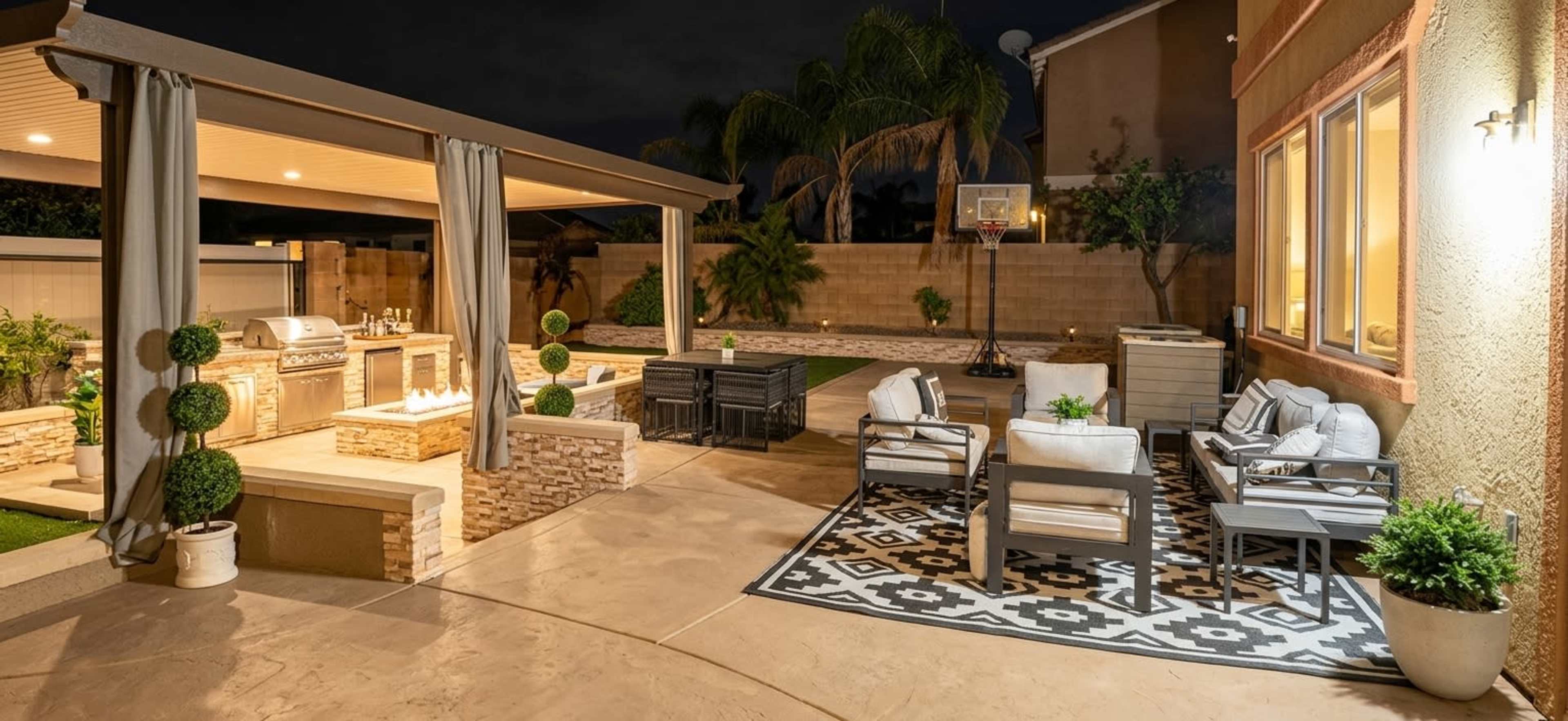 The image shows a well-lit outdoor patio area featuring a seating arrangement, a grill station, and a basketball hoop in the background.