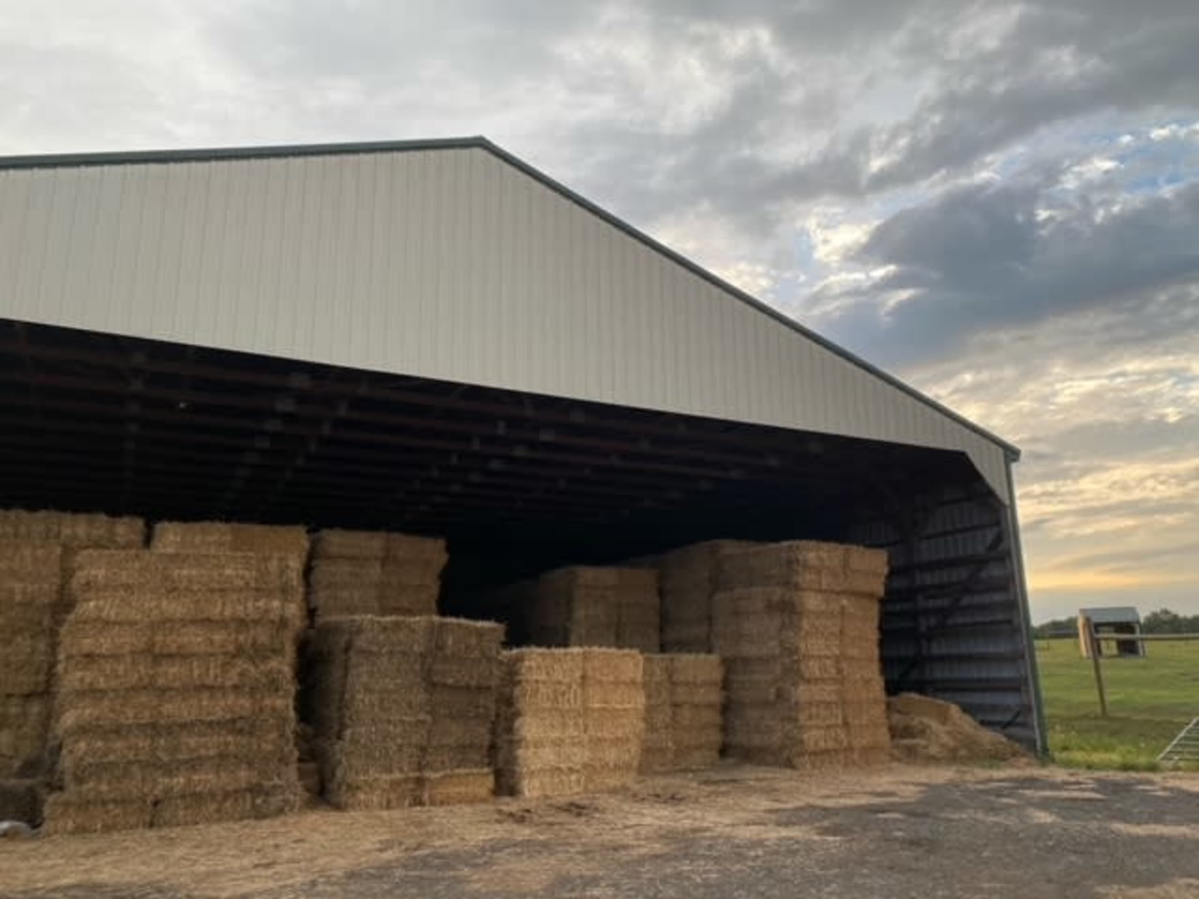 A large barn stores neatly stacked bales of hay inside, with a cloudy sky visible in the background.