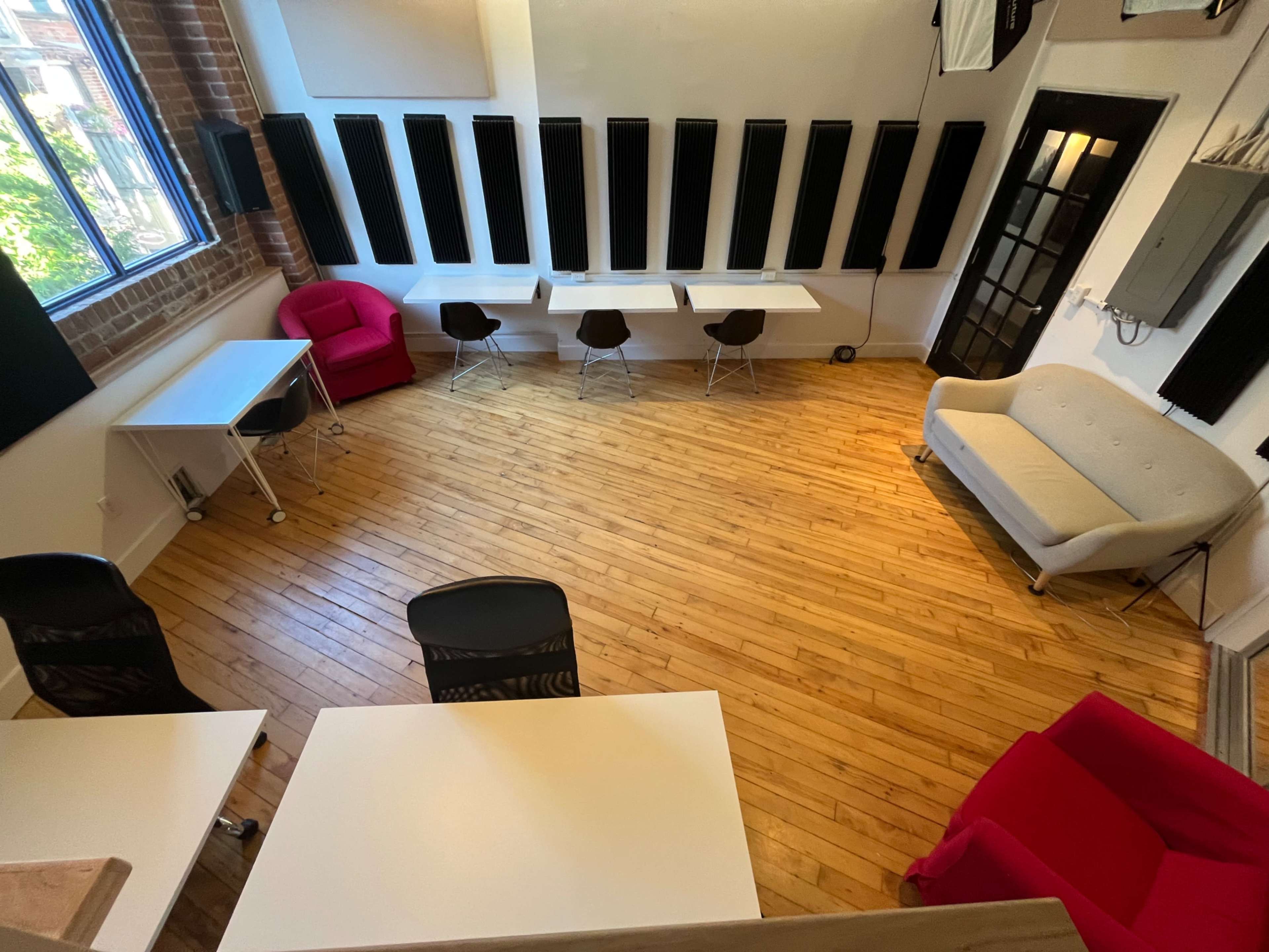 The image shows a spacious room with wooden floors, two red chairs, a gray sofa, and several tables arranged in a modern layout, complemented by soundproofing panels on the walls.