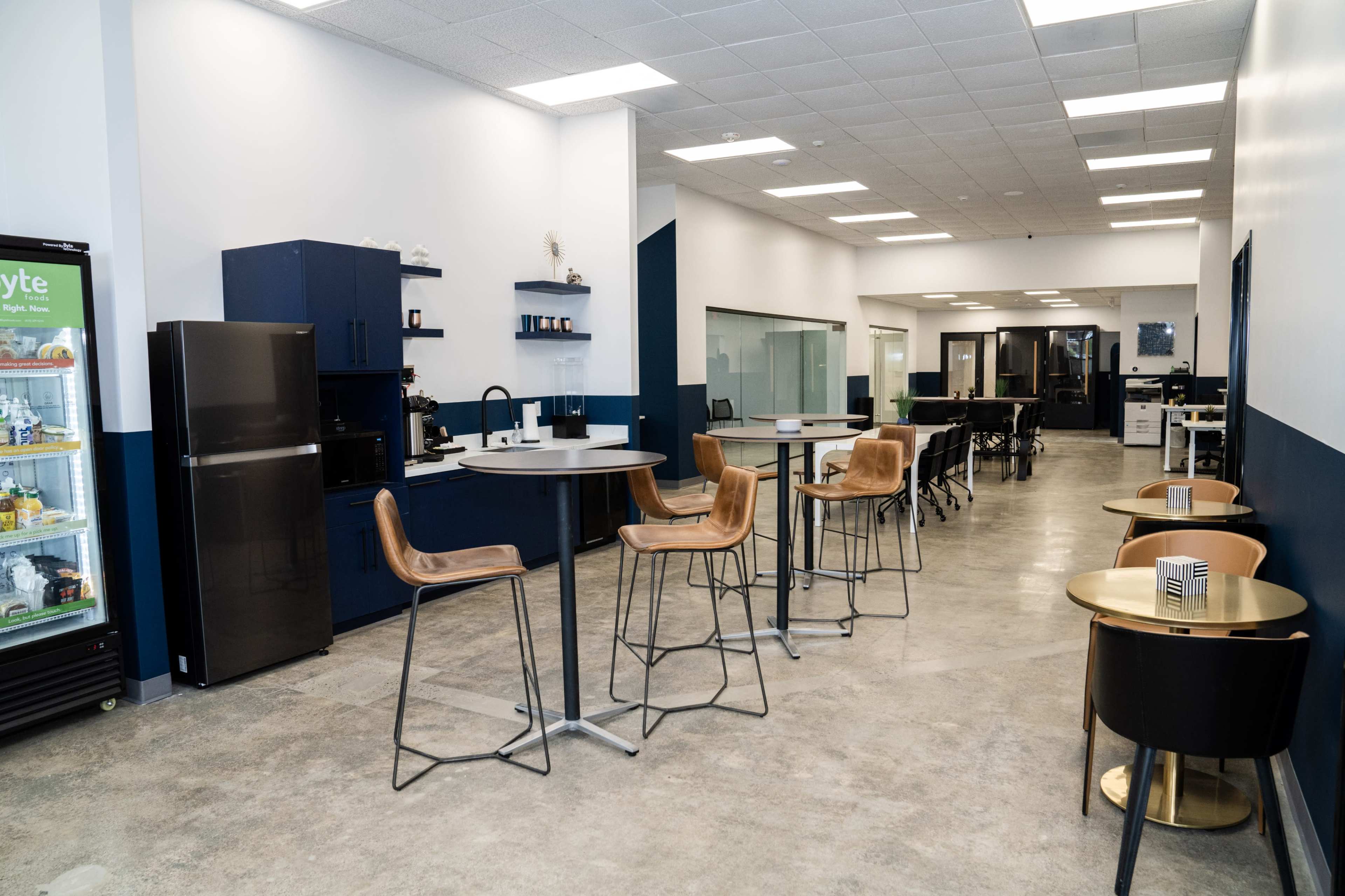 A modern office break area with high tables, bar stools, a fridge, and a coffee station.