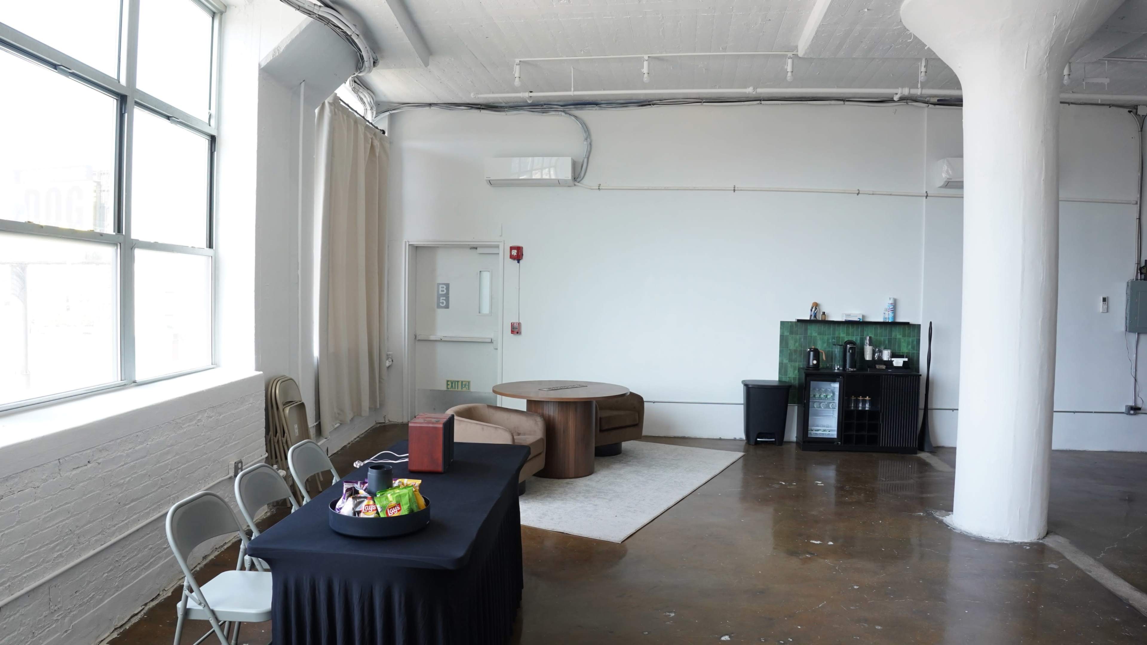 The image shows a spacious, well-lit room featuring a round brown seating area, a black table with snacks, and a small bar-style setup against a white wall.