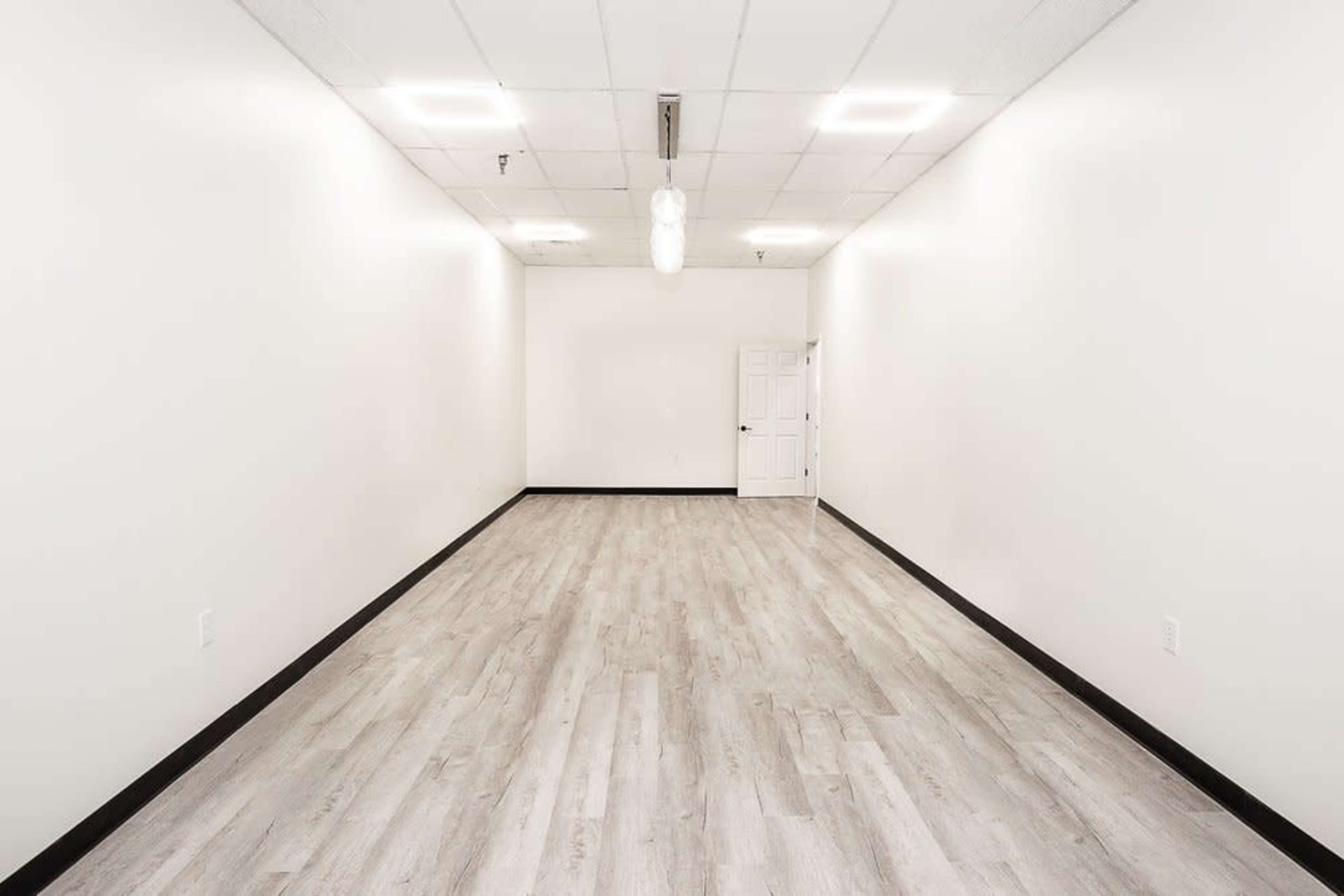 A vacant room with light-colored walls, a wooden floor, and a door at the far end.