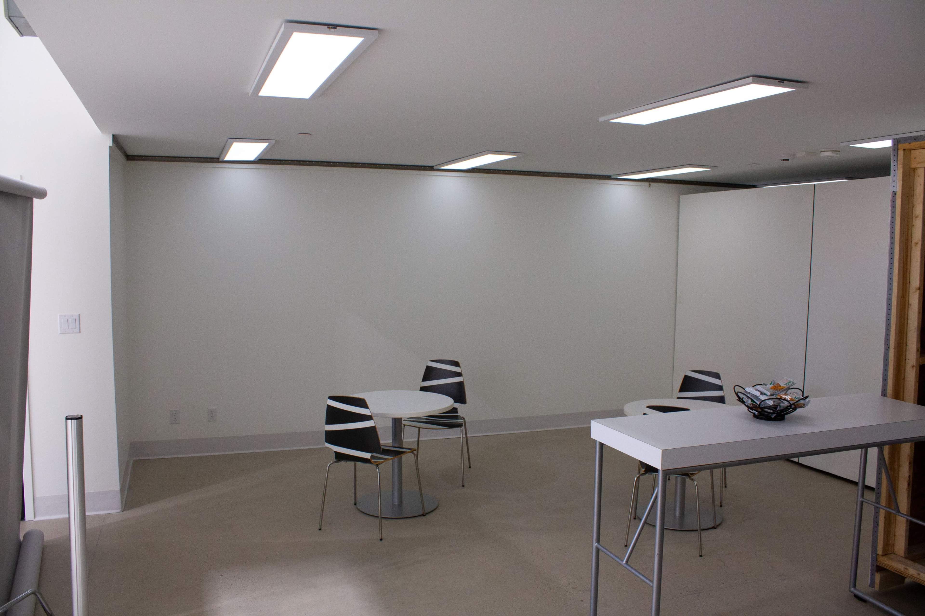The image shows a sparsely furnished room with three chairs and two tables, along with a white countertop and minimal wall decor.
