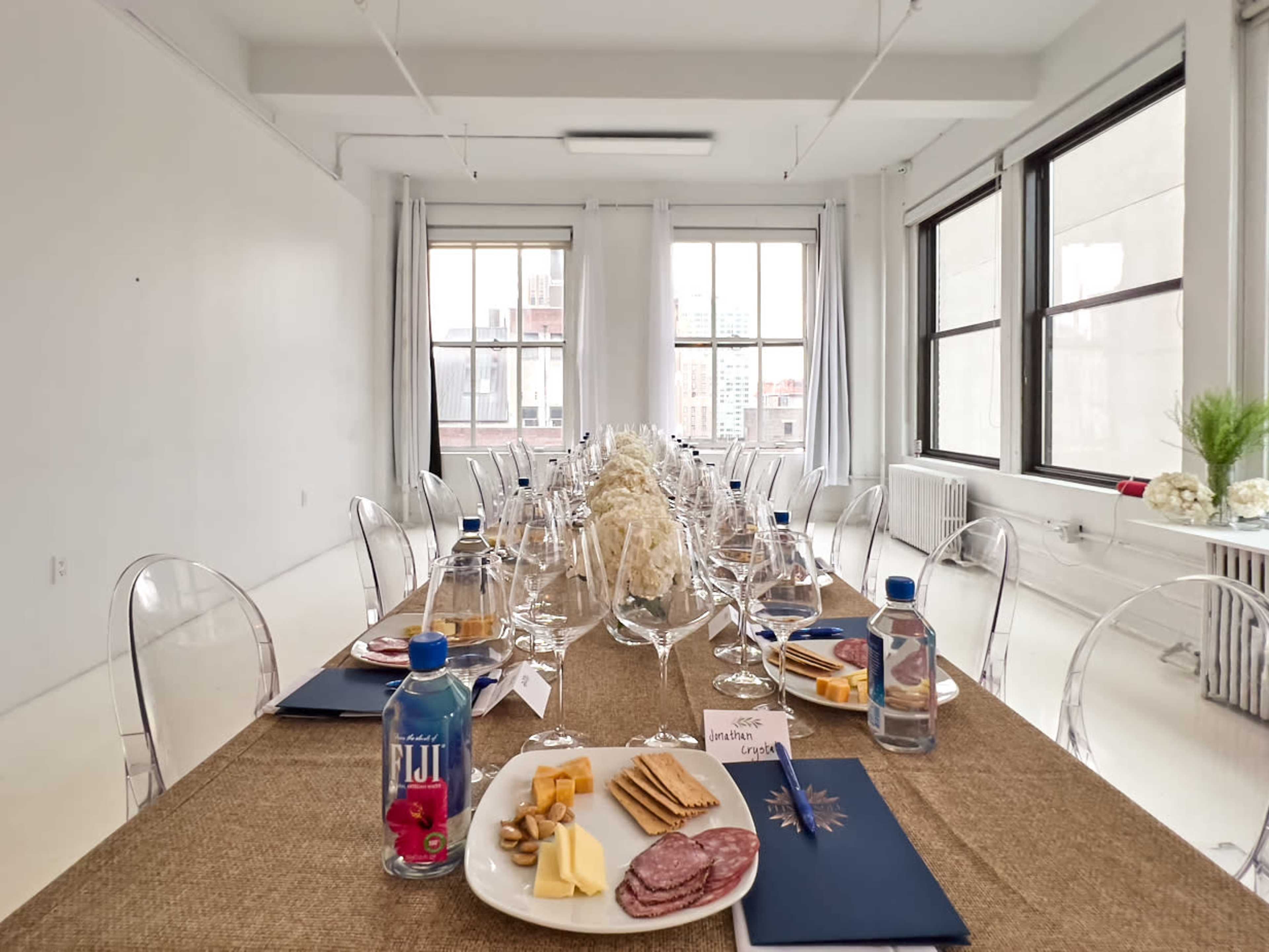 A long dining table is arranged with glasses, plates of cheese, charcuterie, and crackers, alongside water bottles, in a well-lit room featuring large windows.