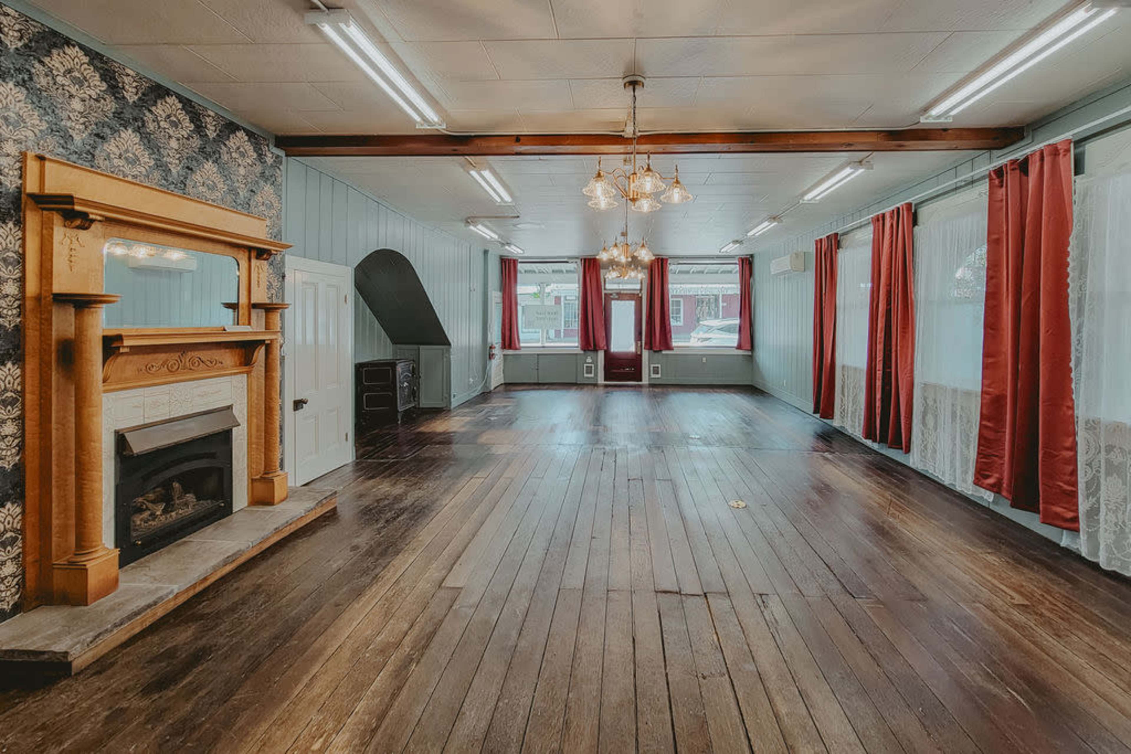 The image shows a spacious interior room with hardwood floors, large windows adorned with red curtains, and a decorative fireplace framed by a wooden mantel.