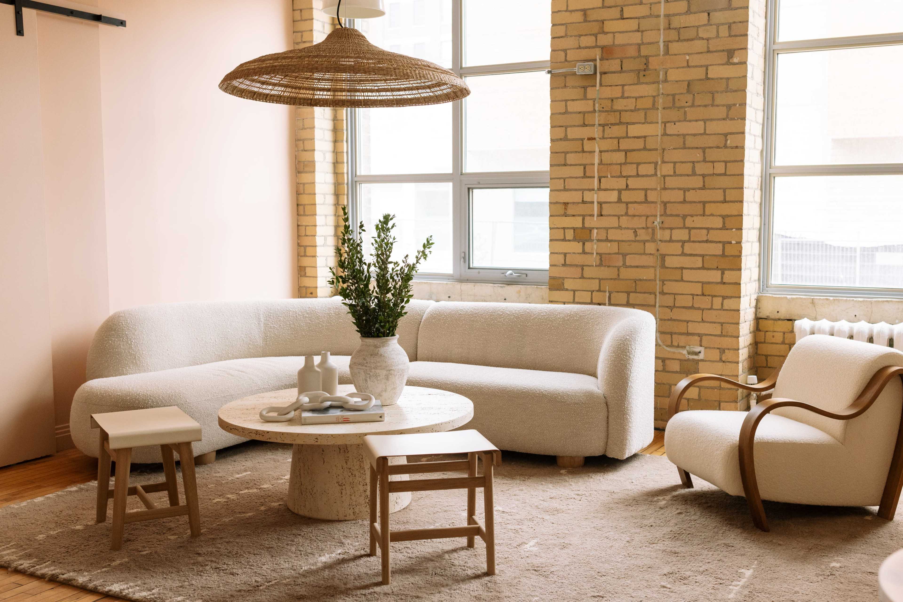 The image shows a modern living room with a curved white sofa, a circular coffee table, and a light-colored armchair, surrounded by large windows and exposed brick walls.