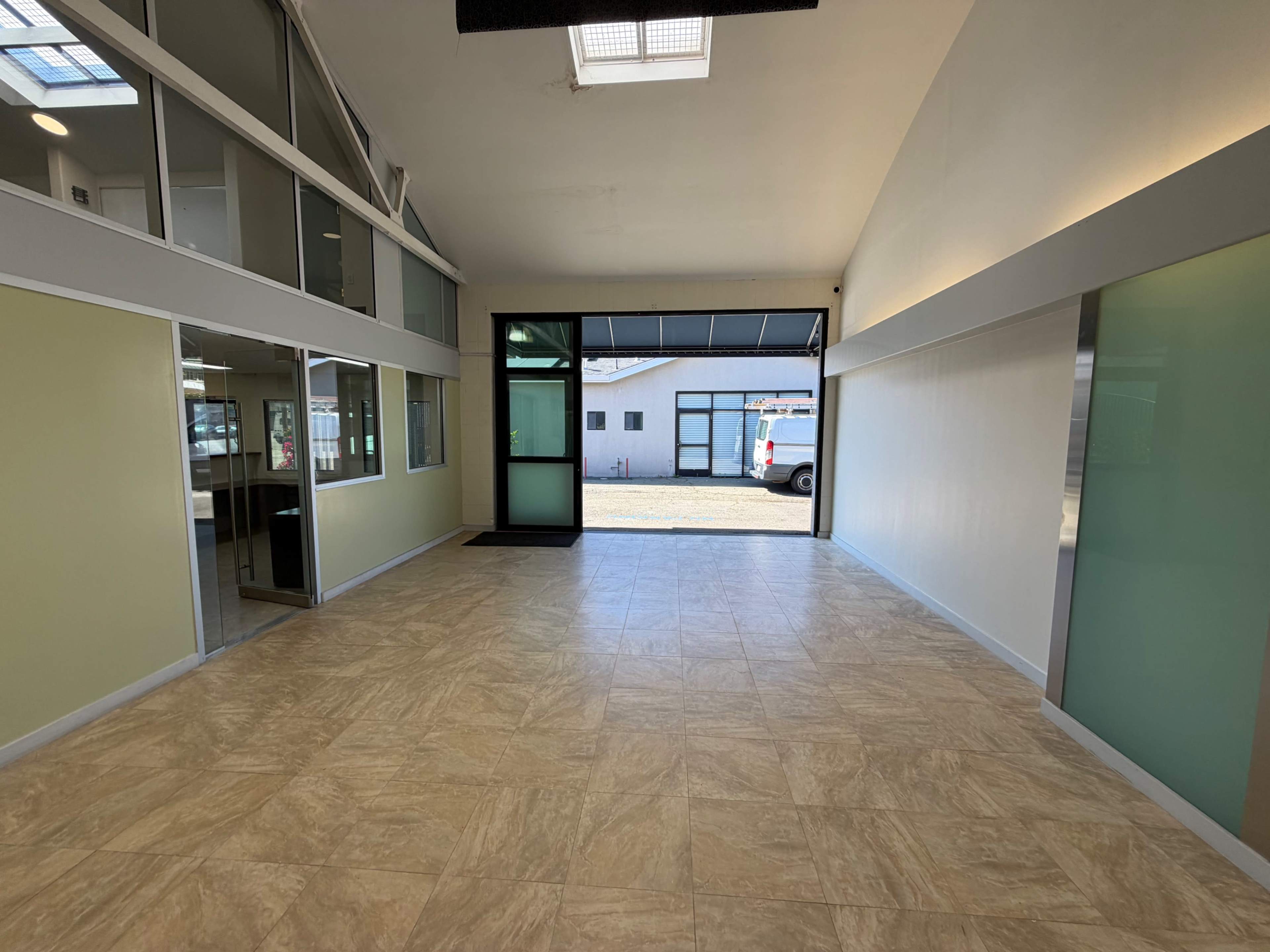 The image shows an empty interior space with a tiled floor, large windows, and a doorway leading to an exterior area.