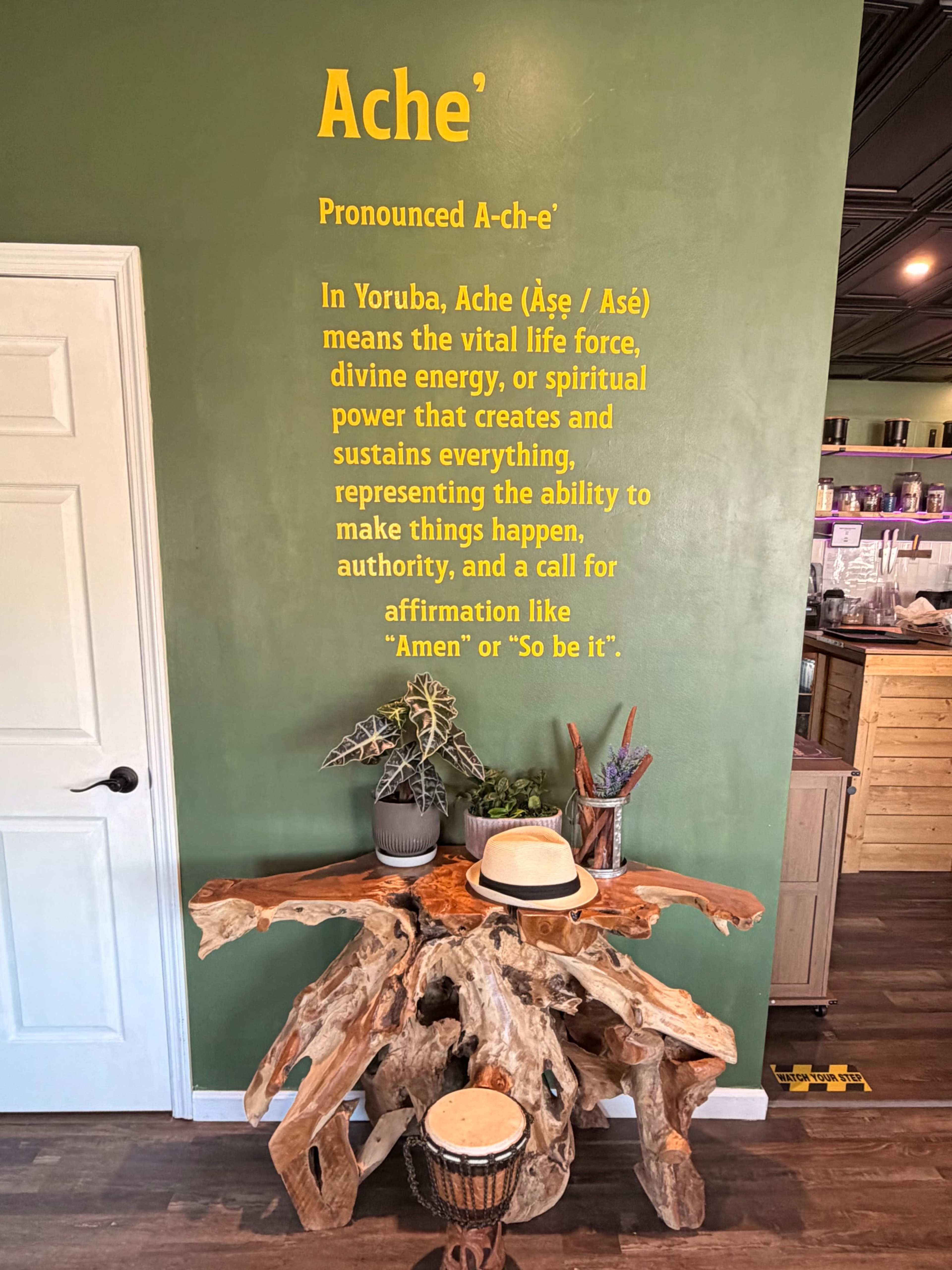 The image features a green wall with text explaining the meaning of "Ache," along with a wooden table and a decorative drum positioned in front.