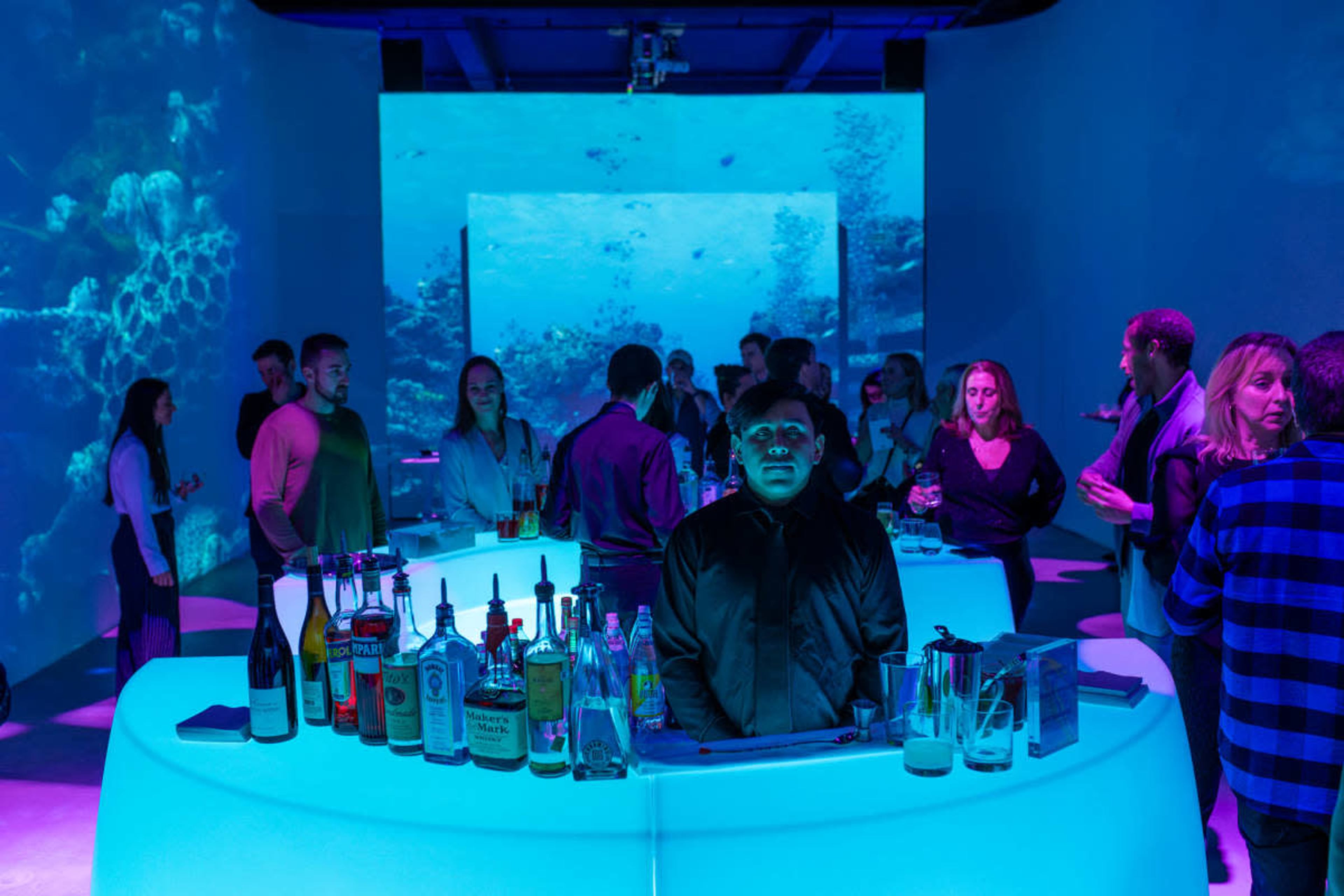 The image shows a vibrant bar scene with patrons socializing, illuminated by colorful projections of underwater visuals in a modern setting.