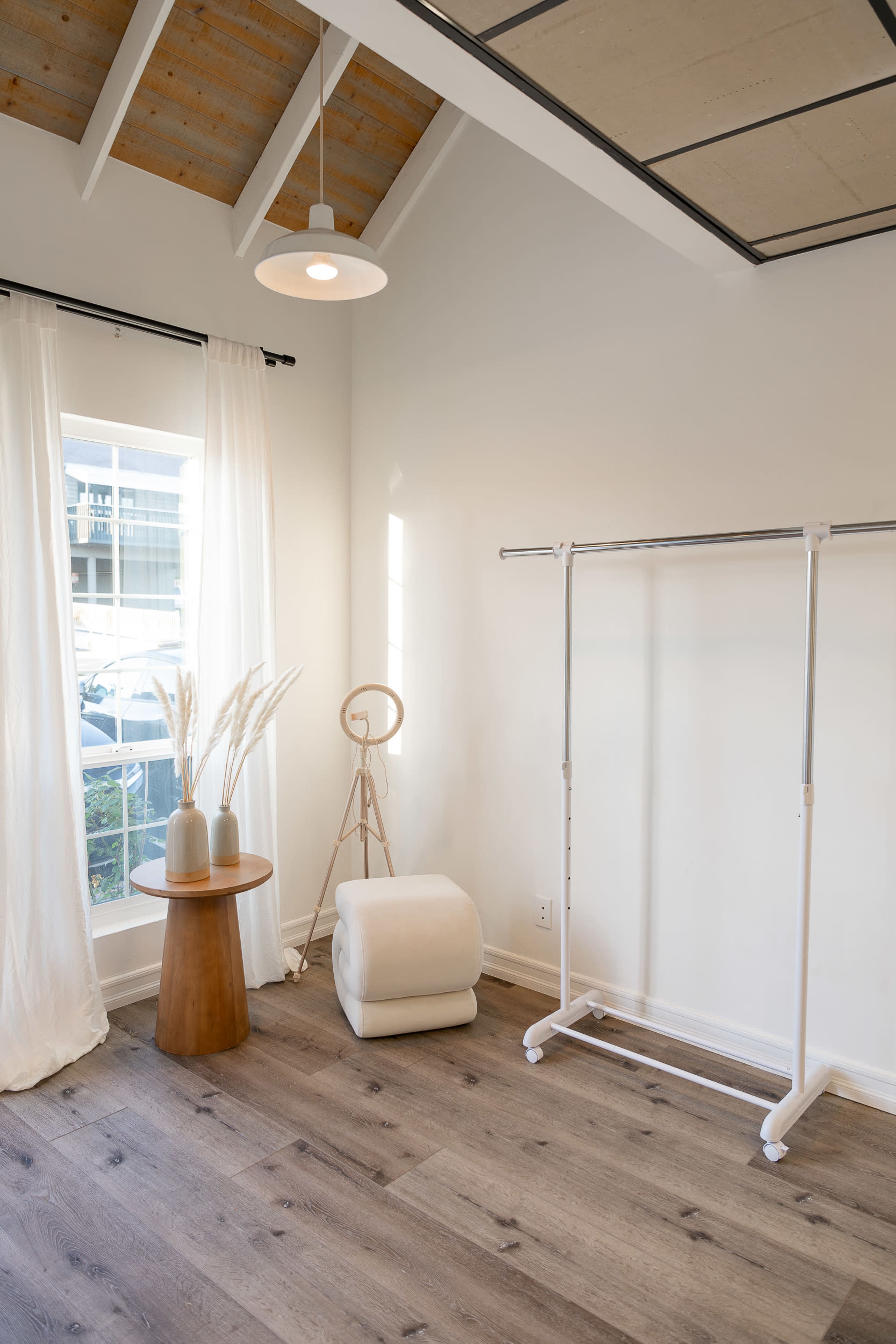 A minimalist room with a wooden table, a white clothing rack, a small white stool, and a large window allowing natural light to enter.
