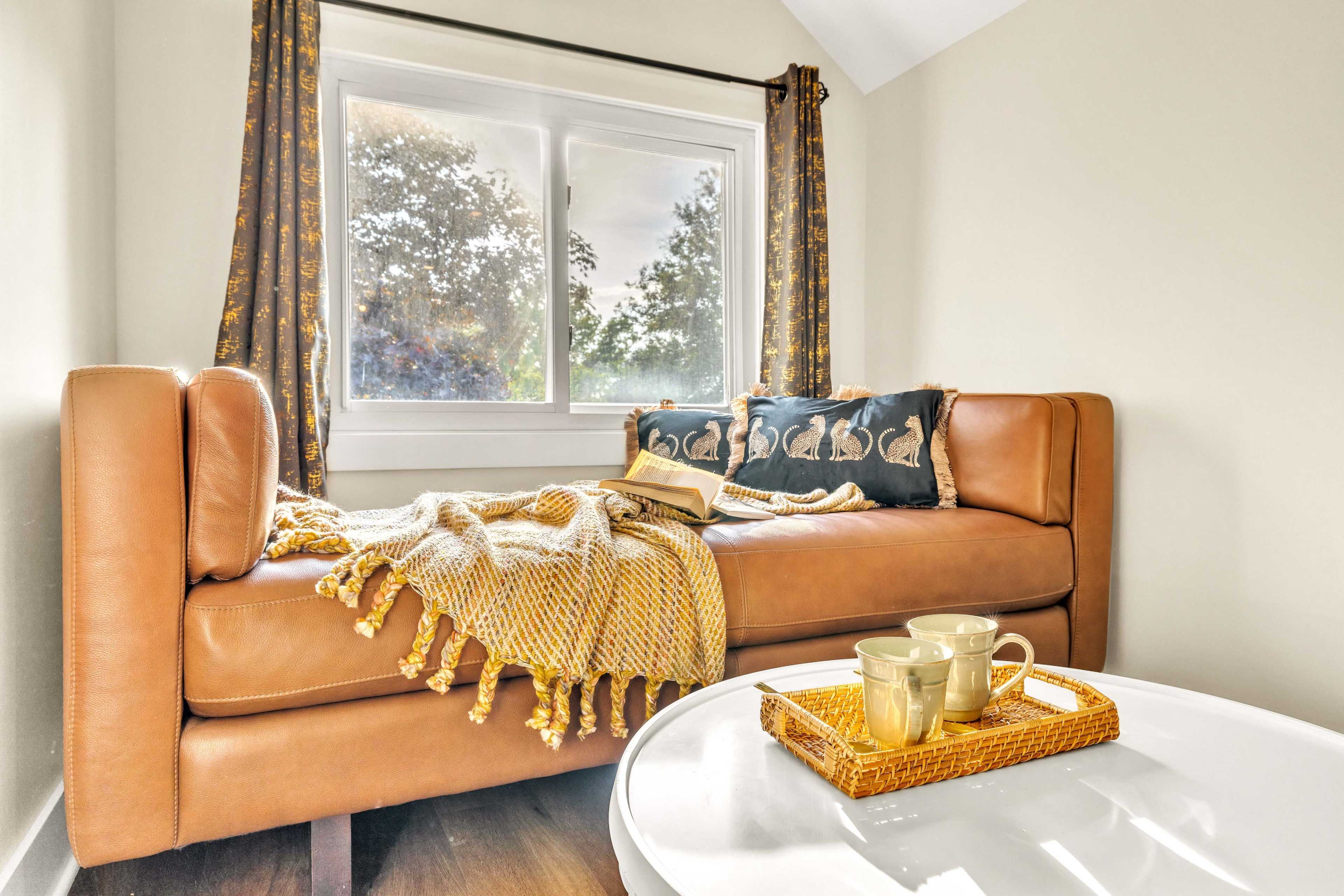 A leather couch with decorative pillows and a throw blanket sits beside a window, with a small table holding a tray and two cups.