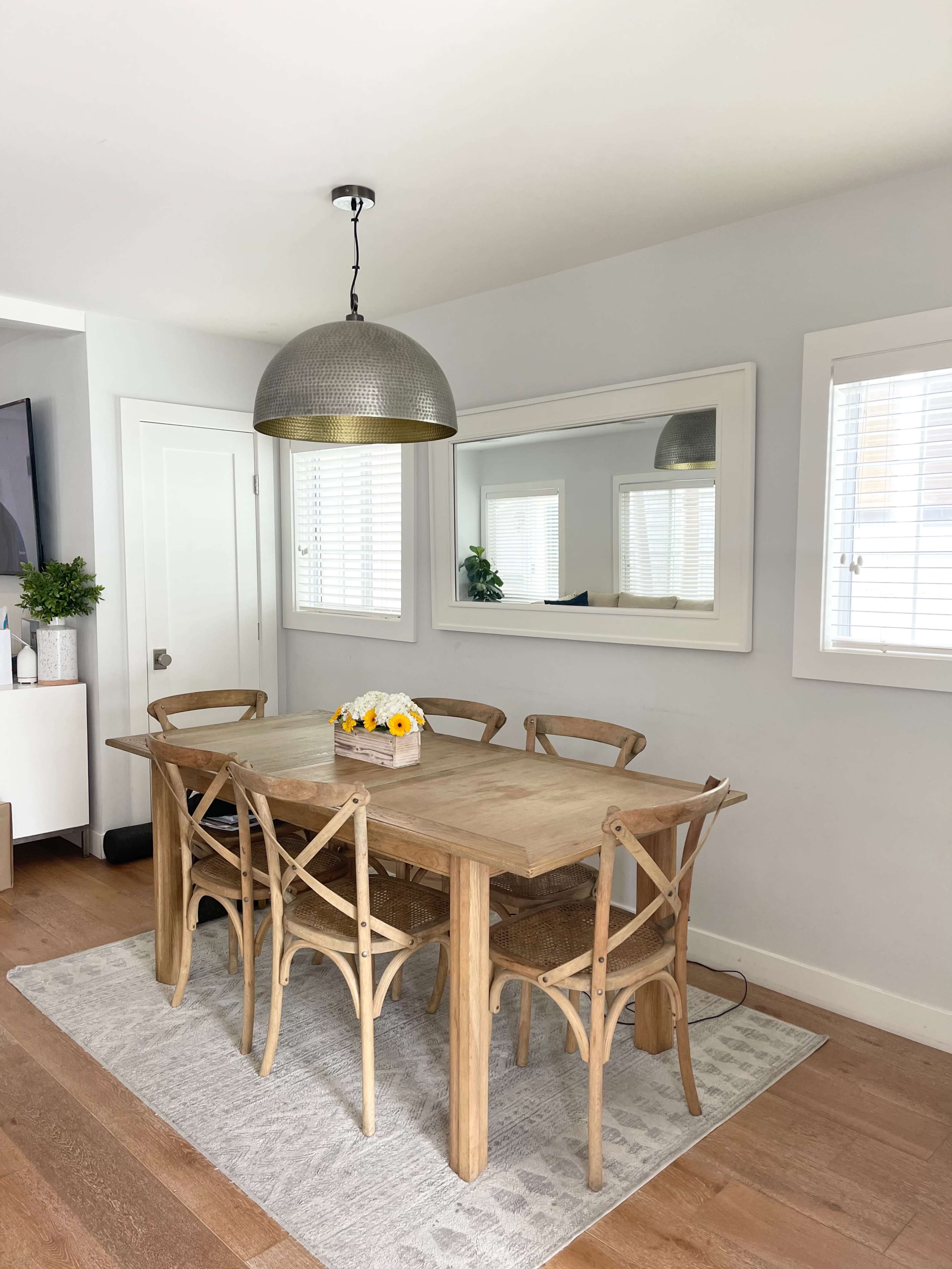 A wooden dining table with six chairs is set in a bright room featuring a large mirror and a modern pendant light.
