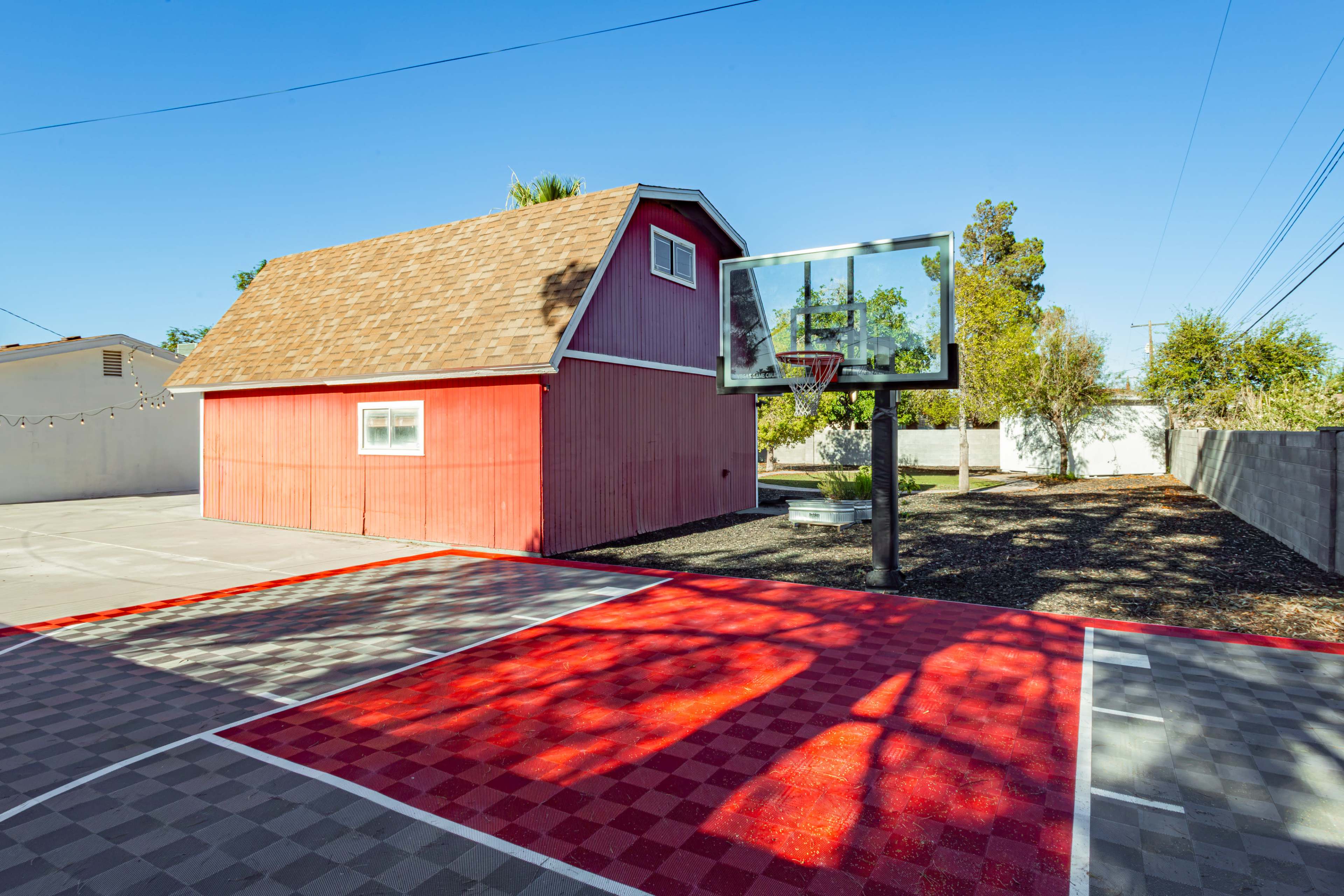 The image shows a red and gray basketball court with a hoop next to a small red shed in a sunny outdoor setting.