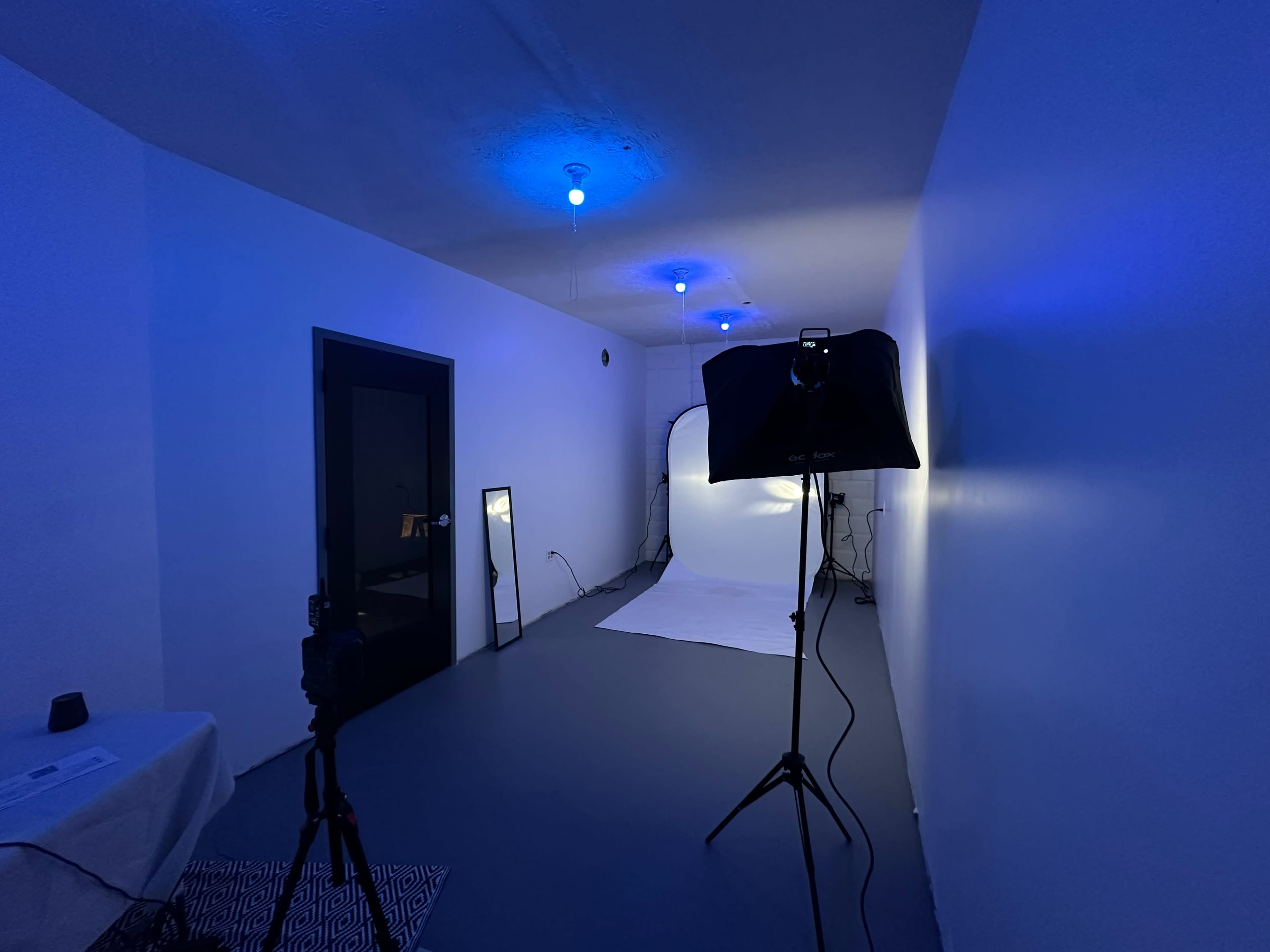 A photography studio with a large white backdrop, a black lighting stand, a mirror, and blue ceiling lights illuminating the room.