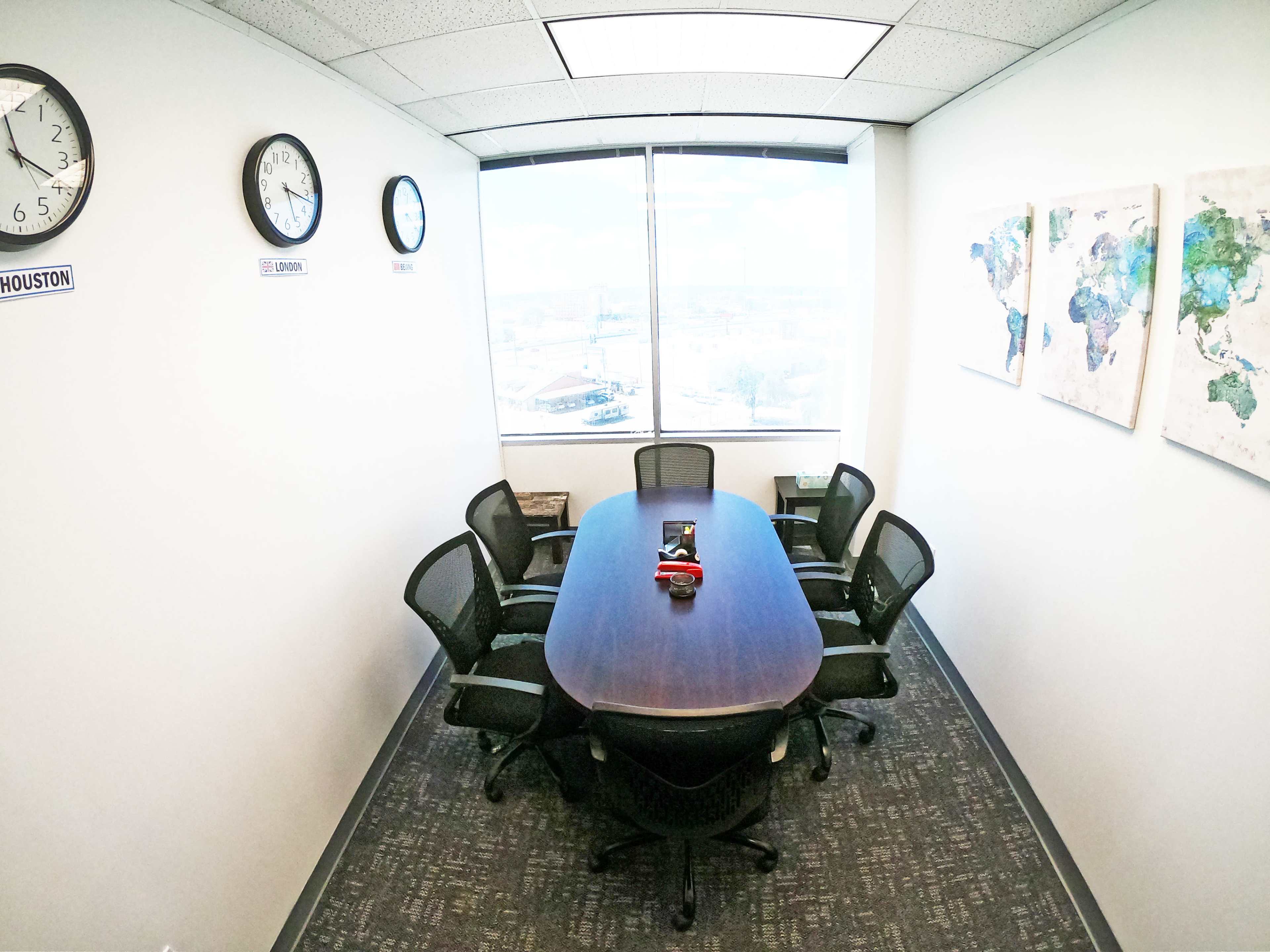 A conference room features a long oval table surrounded by six black chairs, with clocks on the wall displaying different time zones and a world map on the adjacent wall.