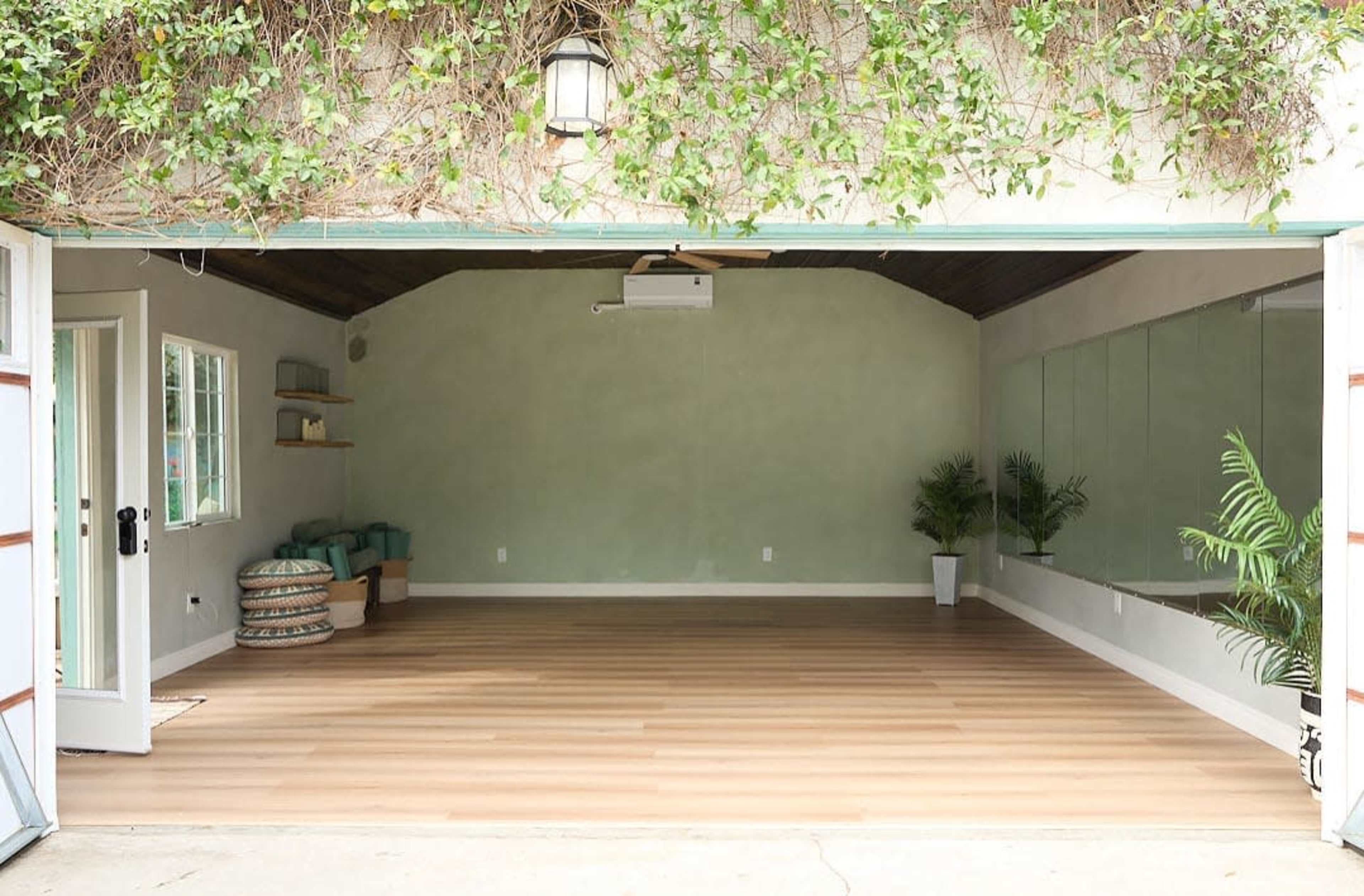 The image shows a spacious, open room with light wooden flooring, a large mirror on one wall, and minimal decor, including a few plants and seating.