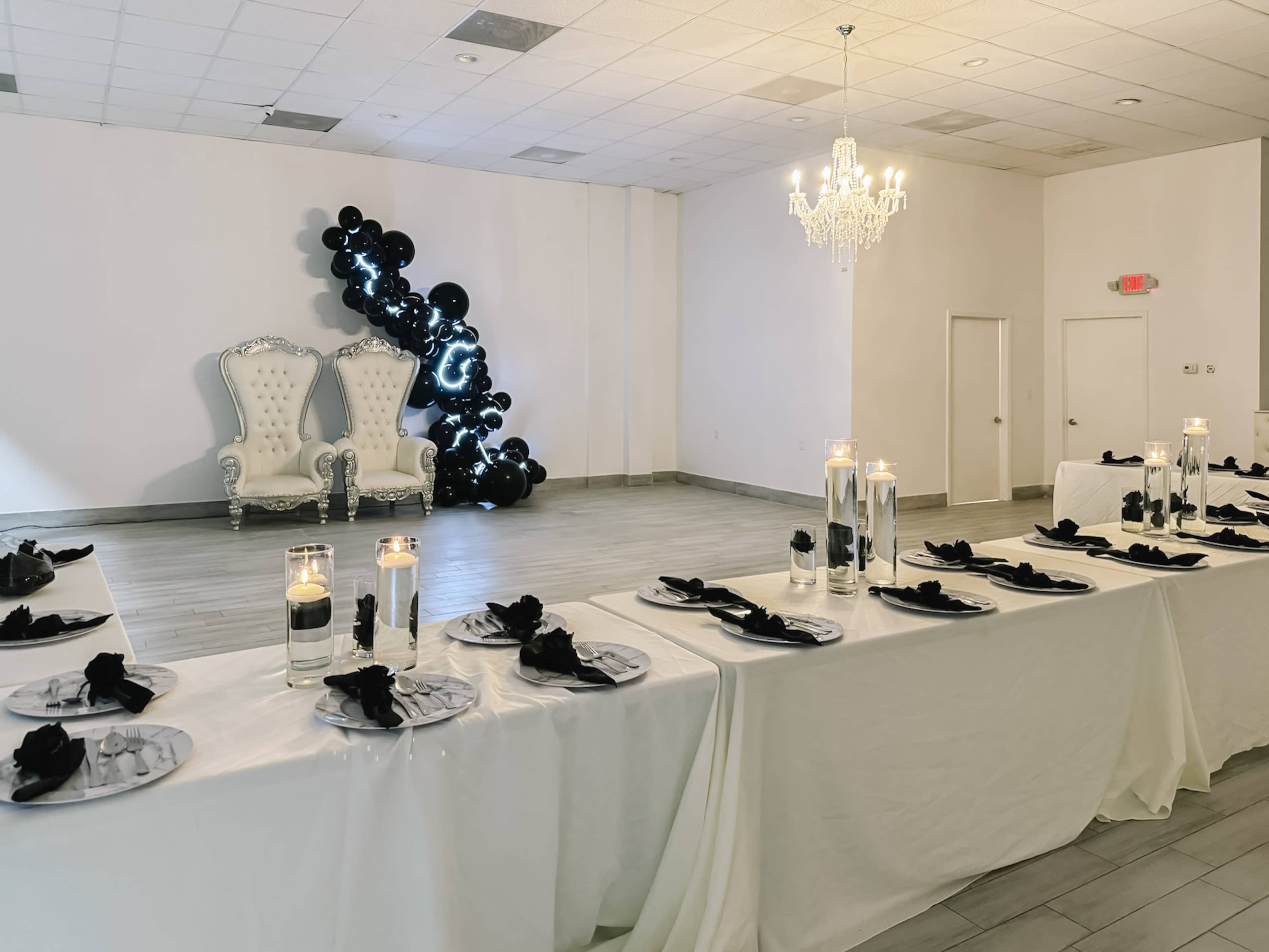 A spacious event room features an elegantly decorated dining table with black napkins and candles, opposite a decorative corner with a white throne and balloon arrangement.