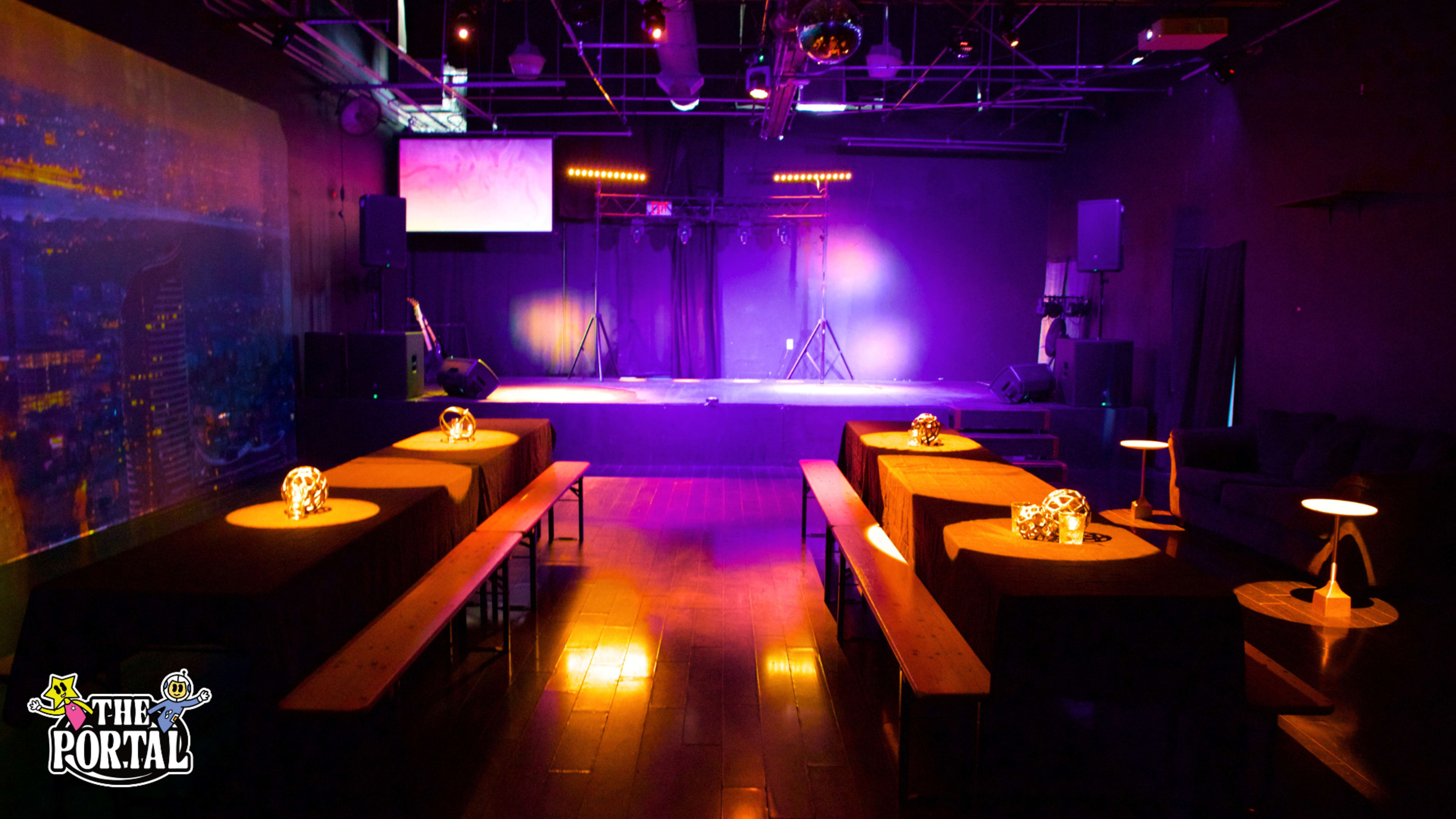 The image shows an empty performance venue with tables arranged in front of a stage, illuminated by purple lighting.