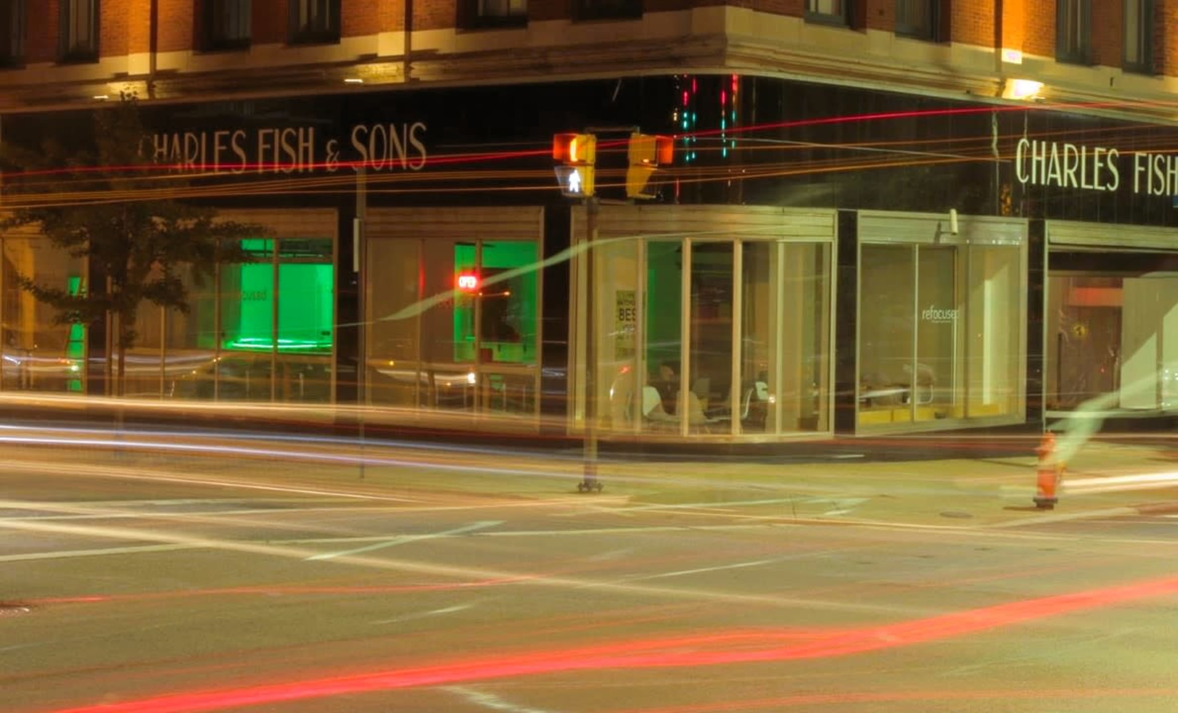 The image depicts a city intersection at night, with light trails from moving vehicles and a brightly lit storefront labeled "CHARLES FISH & SONS."