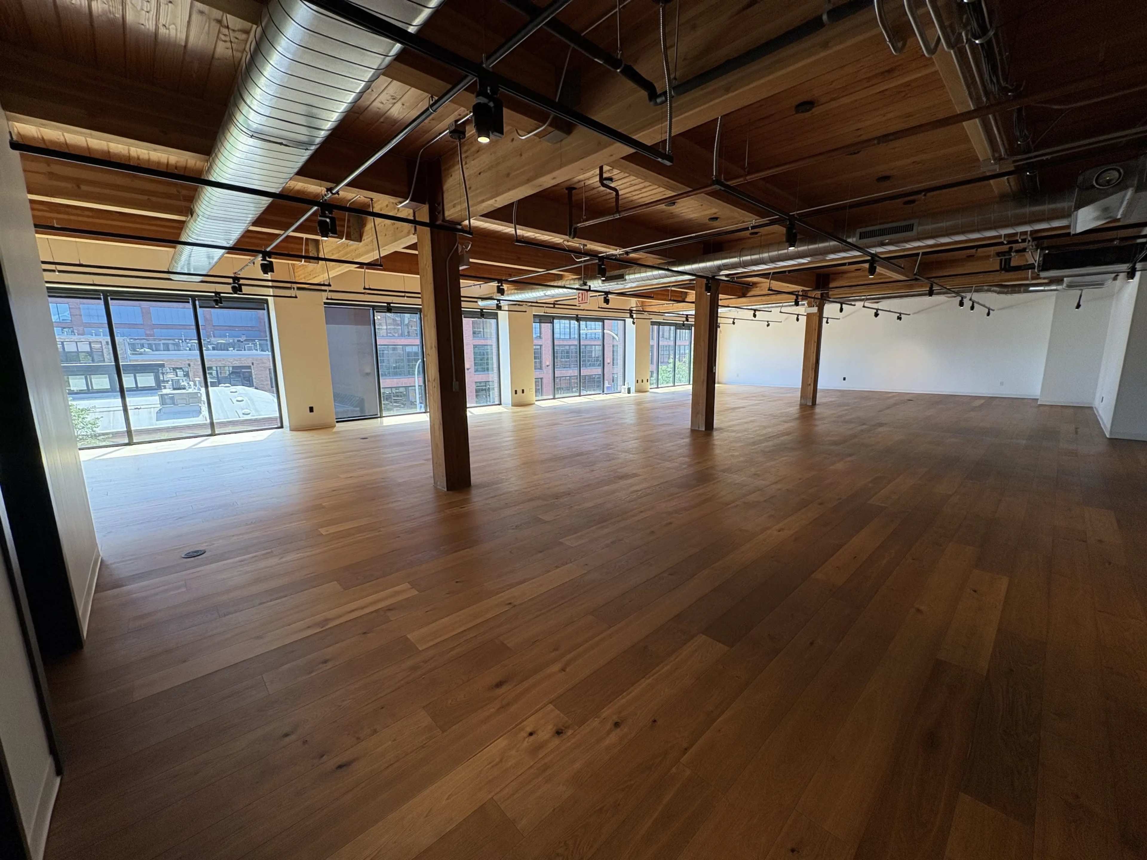 The image shows a spacious, empty interior office space with large windows and wooden flooring.