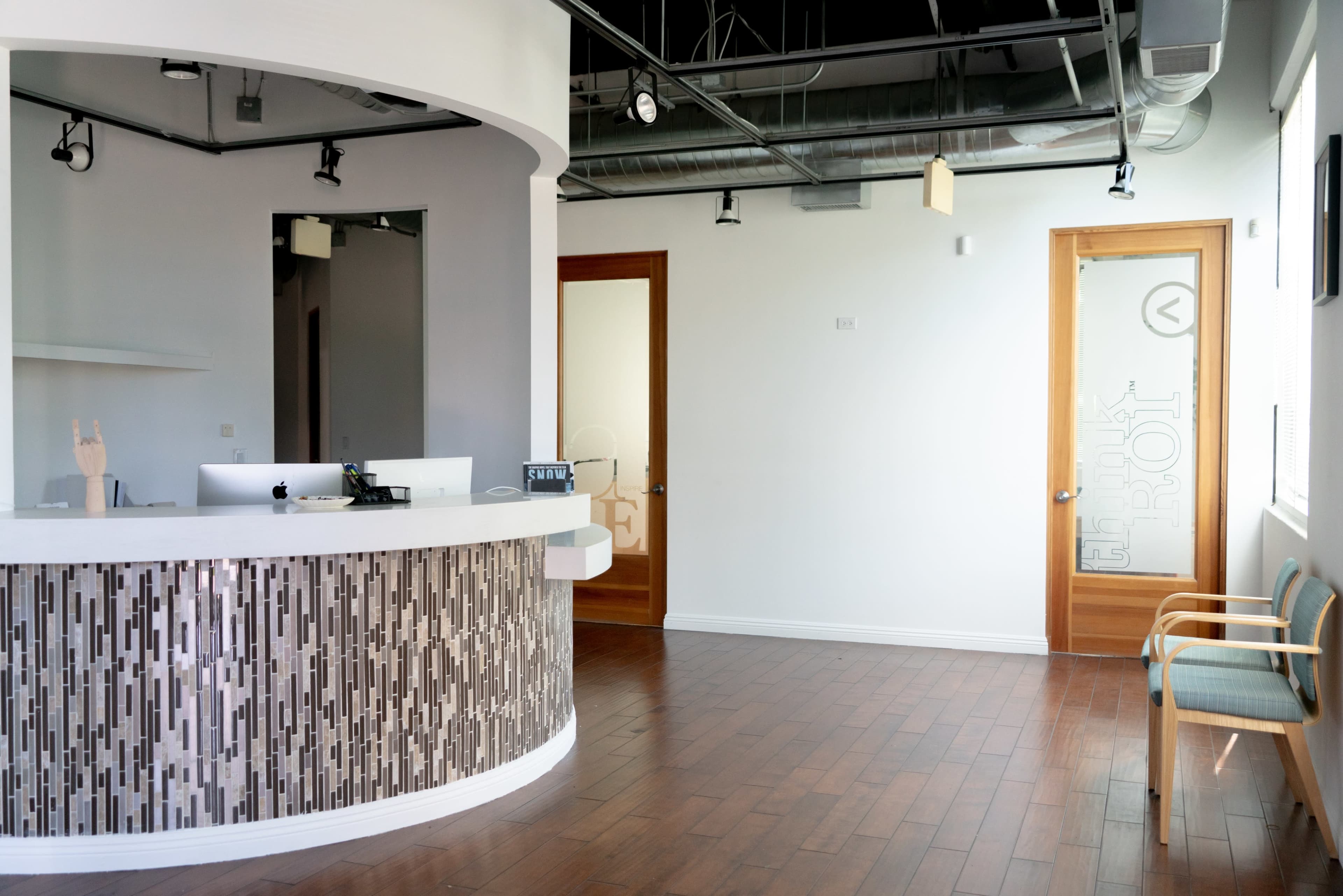 The image shows a modern reception area with a curved front desk, wooden flooring, and a door leading to another room.