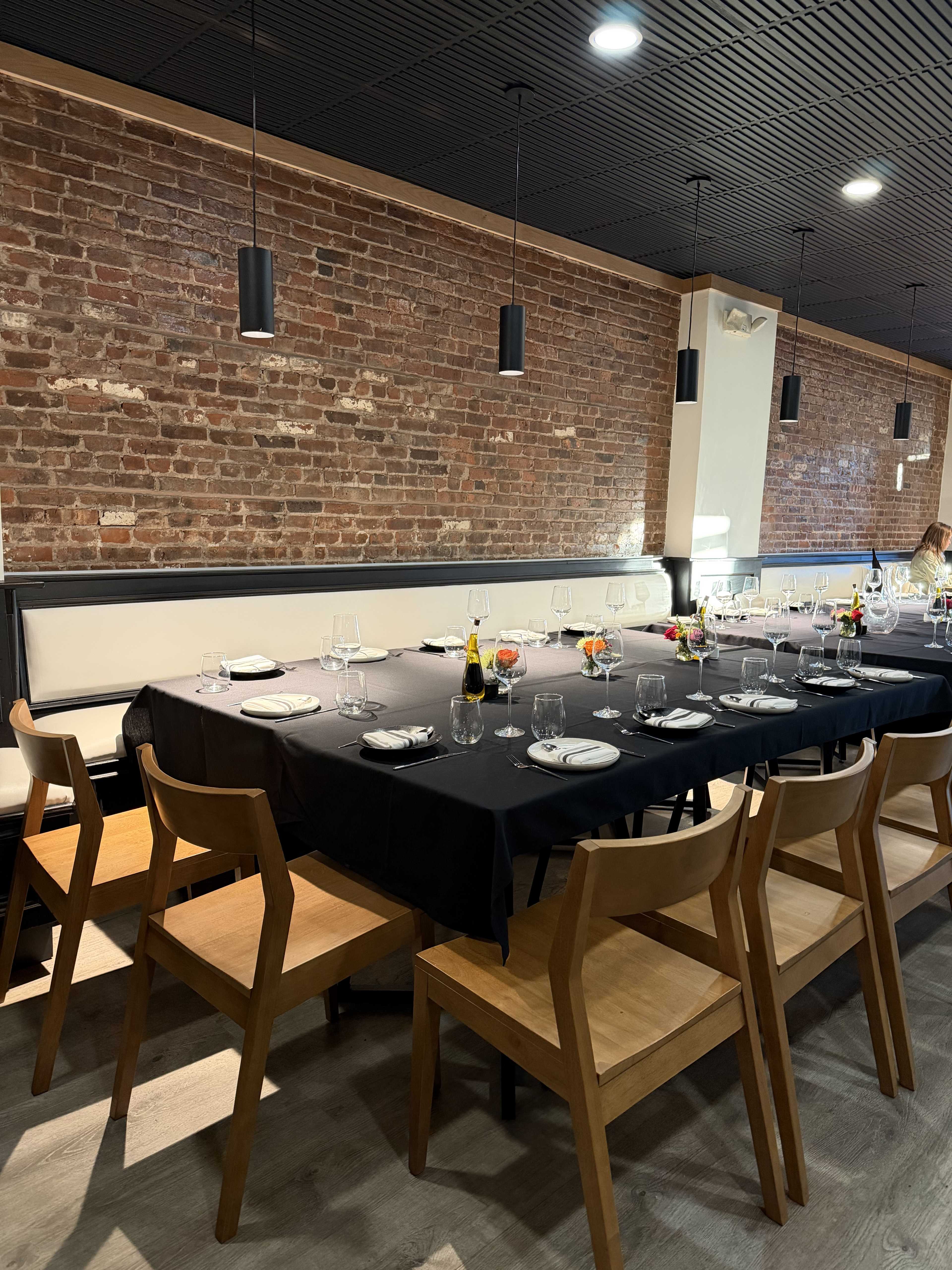 The image shows a dining area with a large black table set for a meal, surrounded by wooden chairs, and featuring a rustic brick wall in the background.