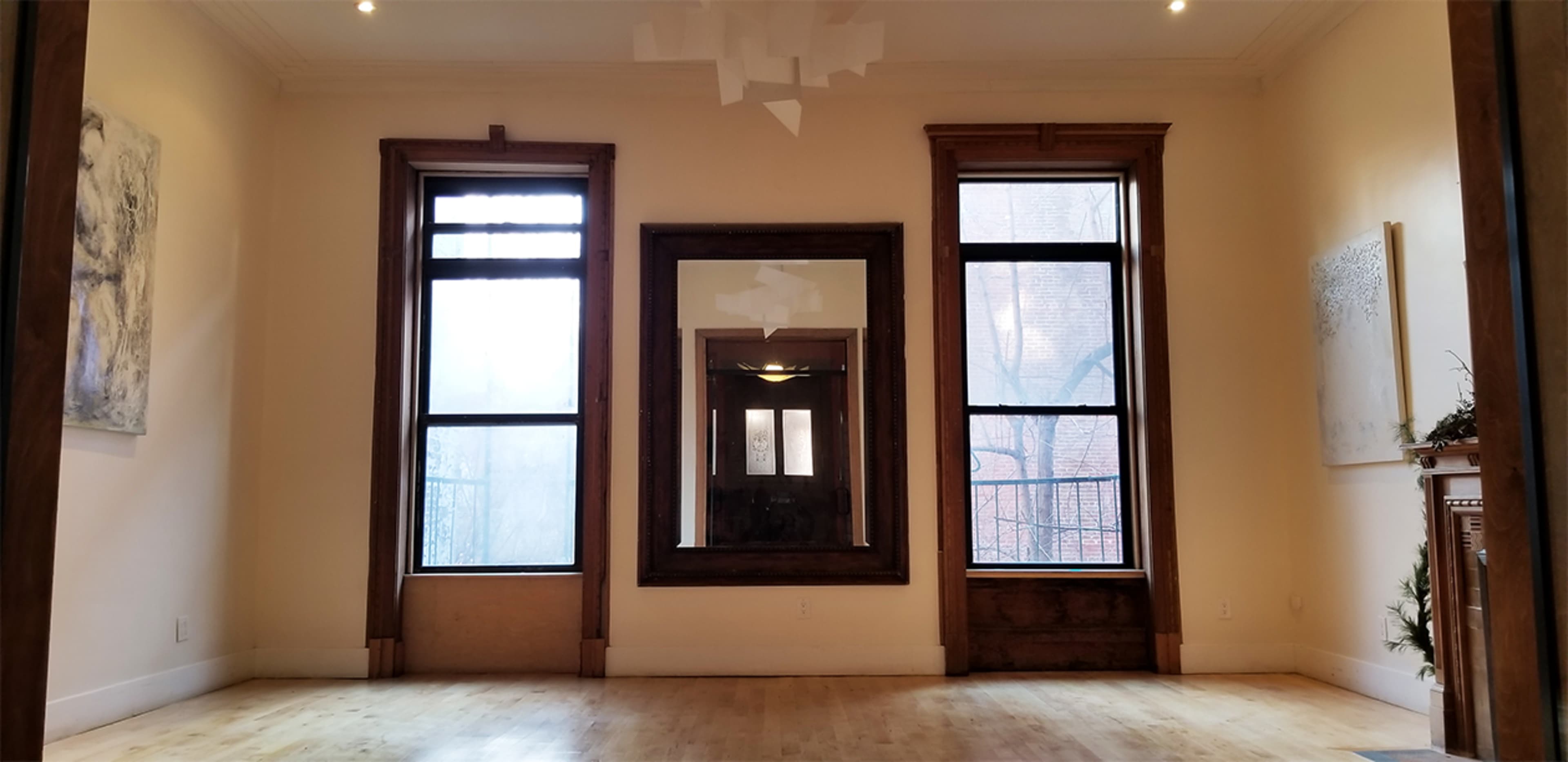The image shows a spacious, empty room with three windows and a large mirror framed in wood, highlighting the wooden trim and light-colored walls.