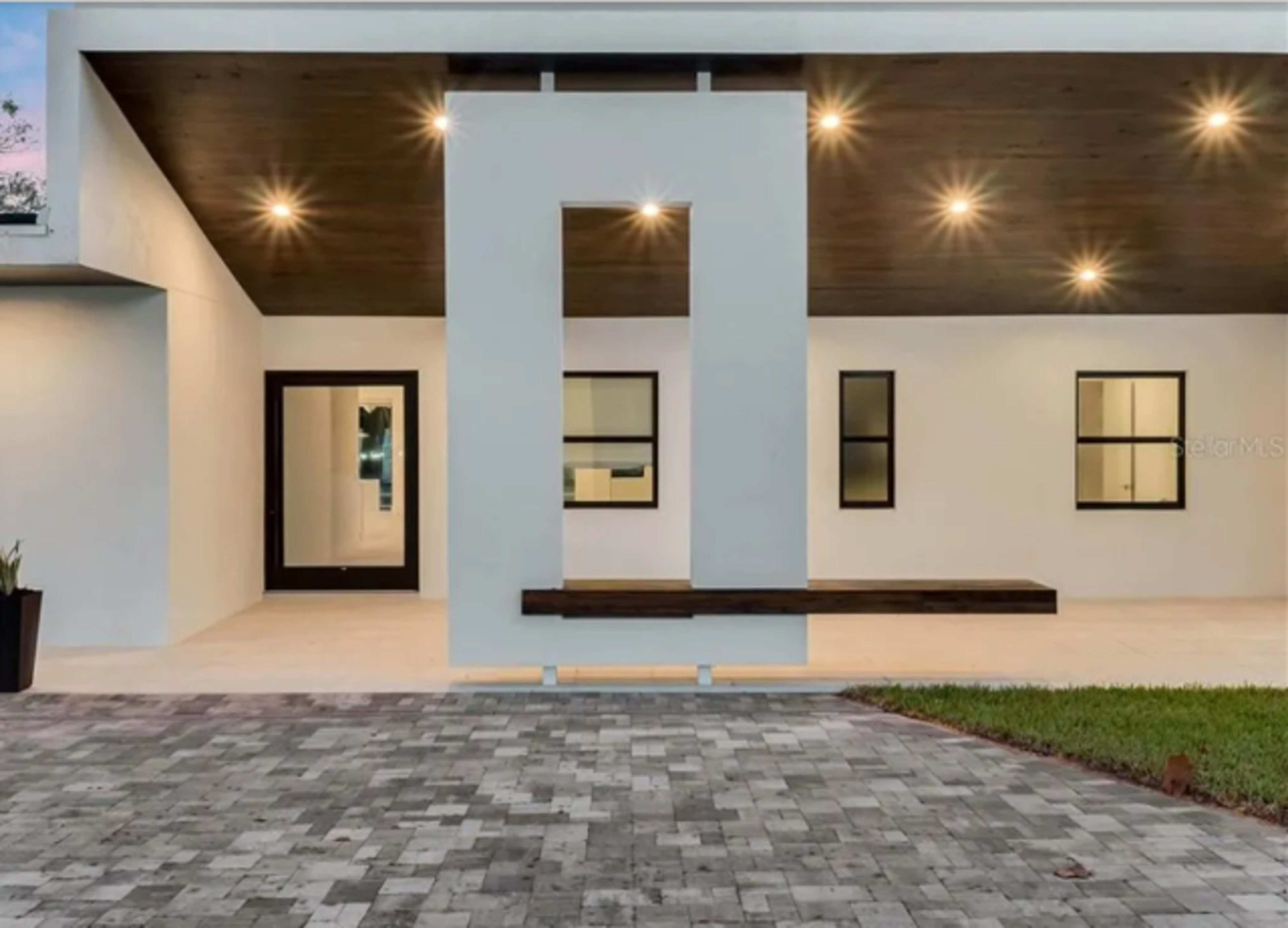 The image shows a modern residential building with a flat roof, a minimalistic entrance featuring a geometric design, and a paved pathway leading up to the door.