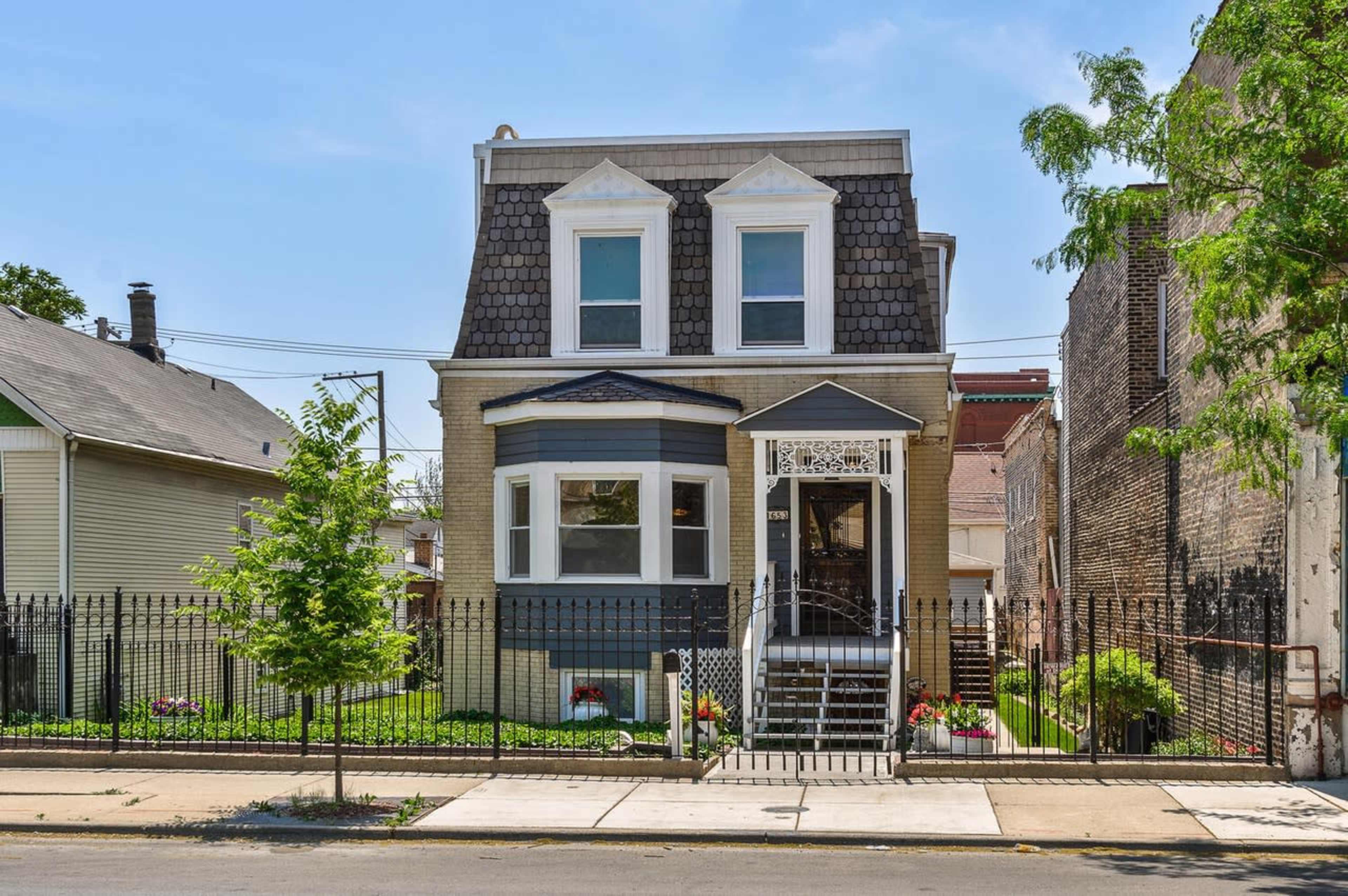 A two-story house with a sloped roof, front porch, and a surrounding wrought-iron fence is situated on a tree-lined street.