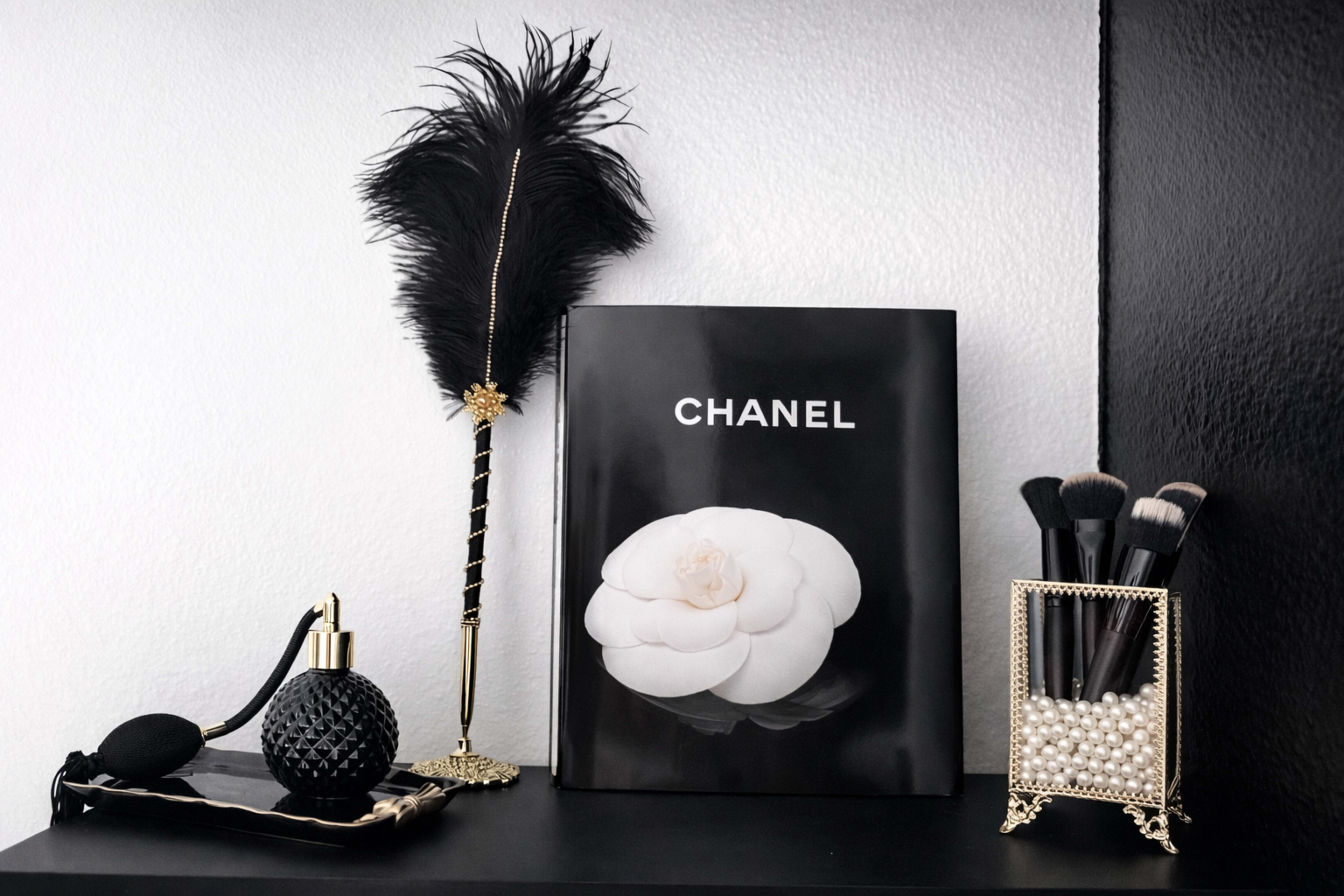 The image shows a stylish shelf featuring a Chanel book, a black perfume bottle, a feather pen, and a holder containing makeup brushes.