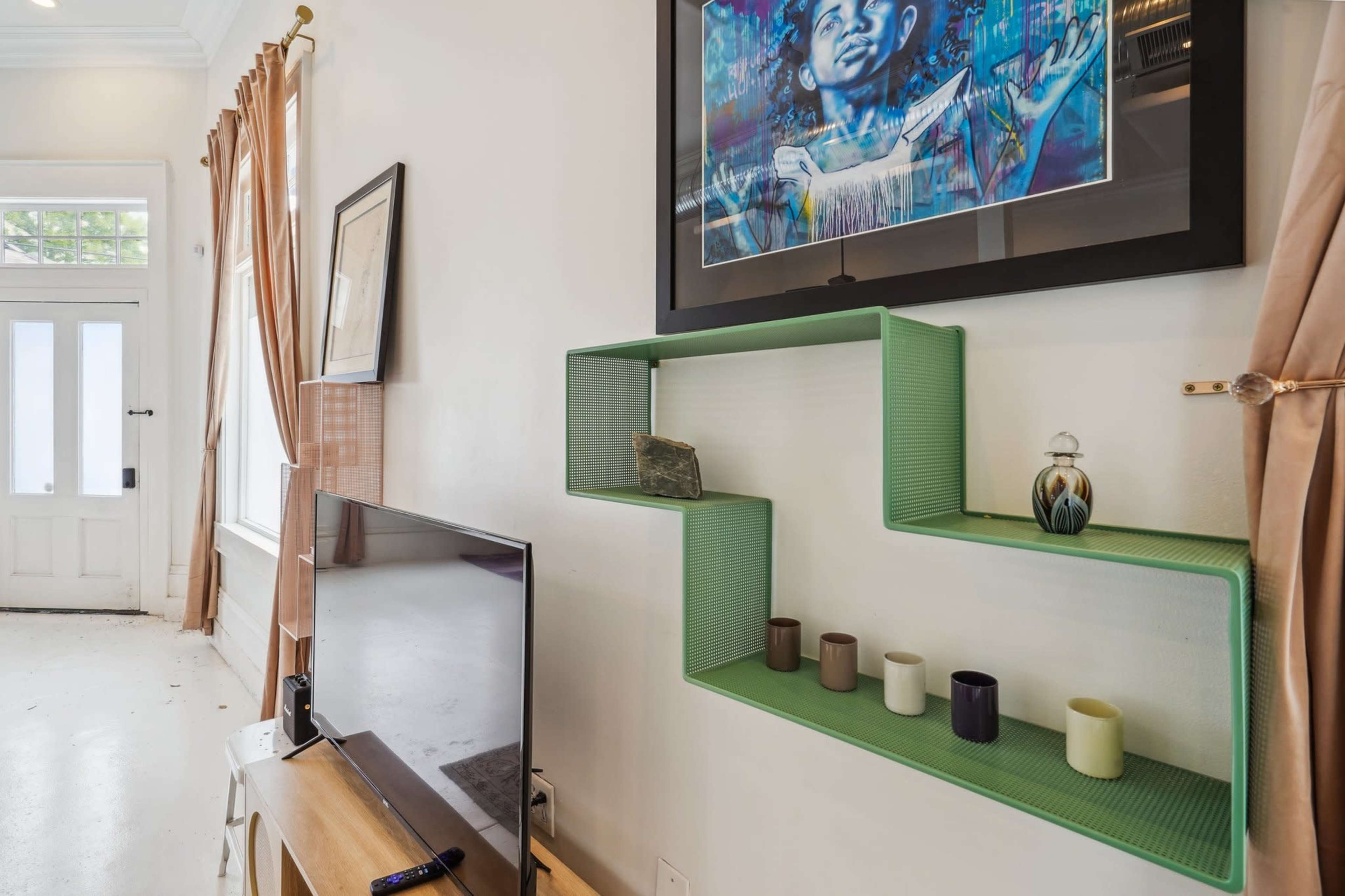 A wall-mounted green shelf displays decorative items next to a television in a brightly lit room with large windows and curtains.