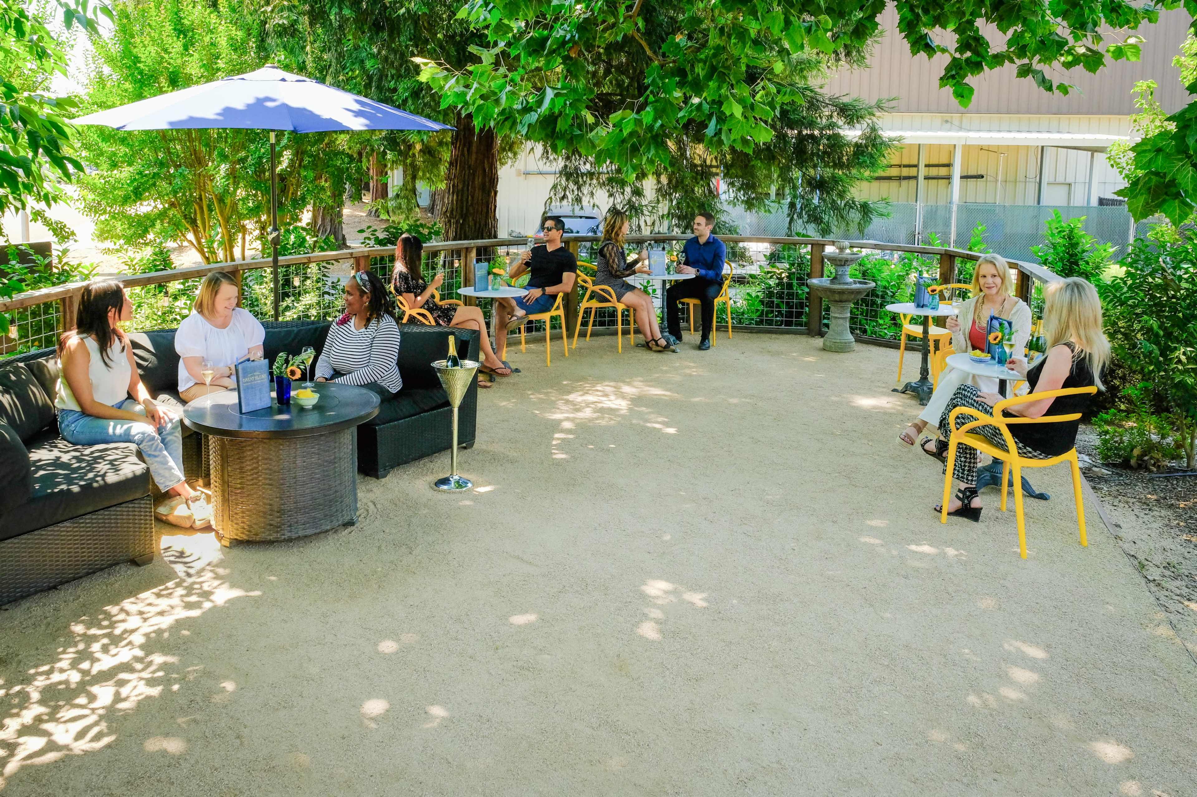 A group of people is seated in a shaded outdoor area, socializing around tables with drinks and snacks.