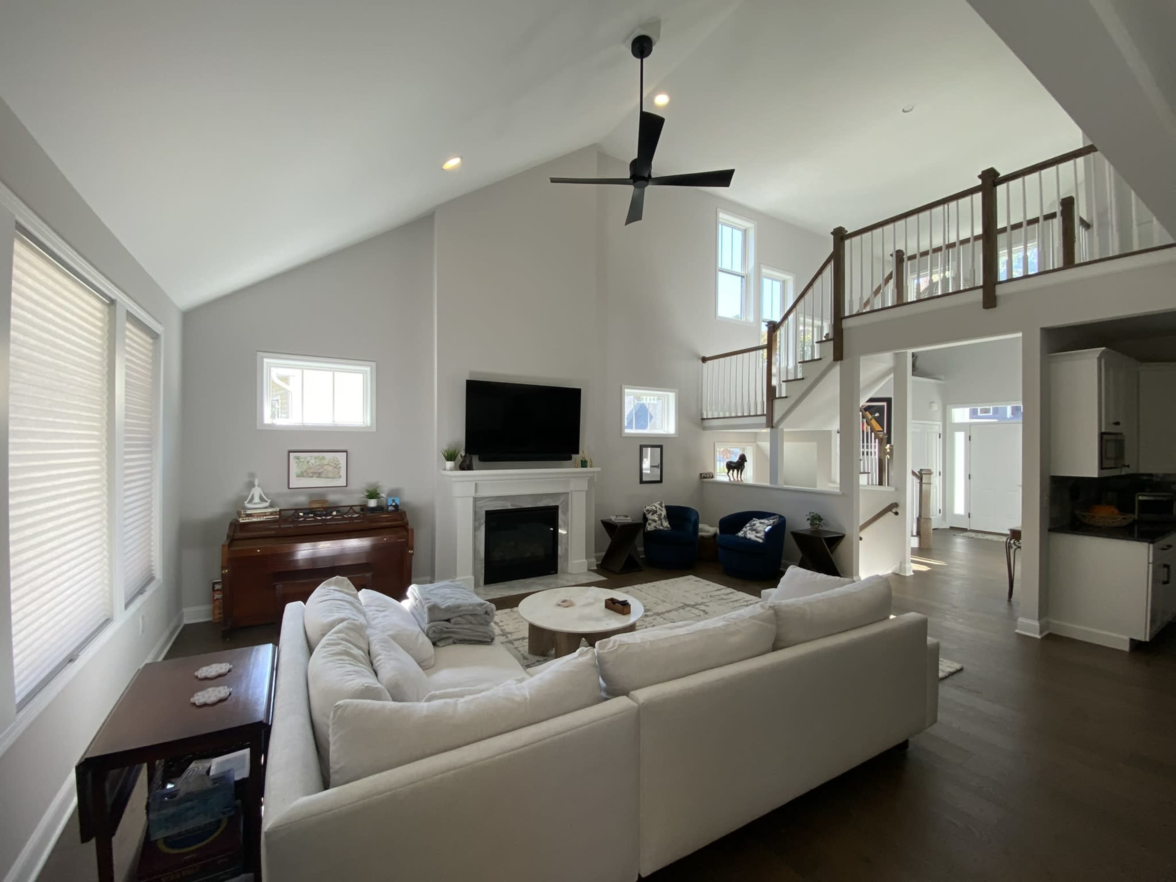 A spacious living room features a white sectional sofa, a large television above a fireplace, and wooden stairs leading to an upper level.