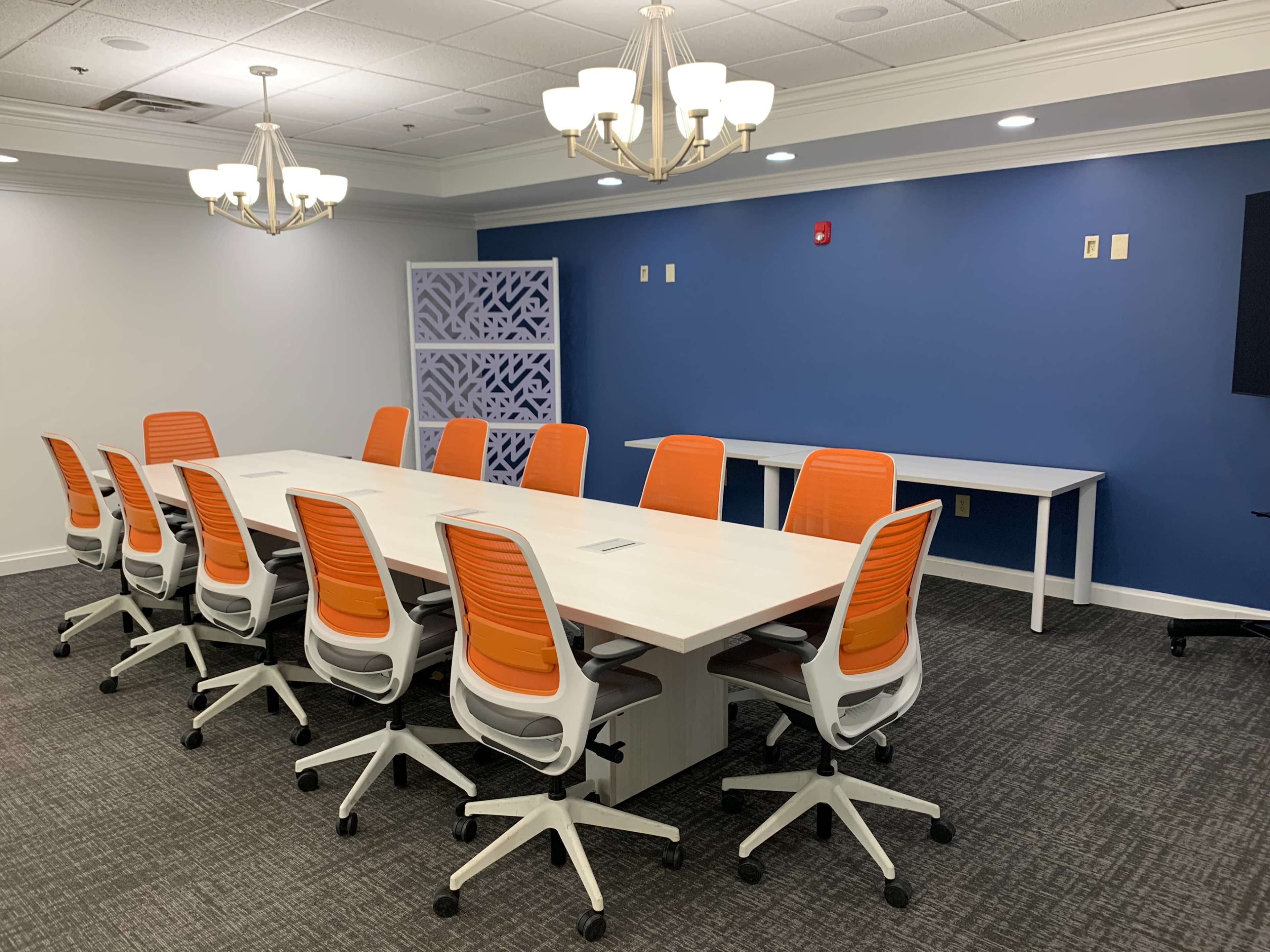 A modern conference room with a long white table surrounded by orange and white chairs, a blue accent wall, and a side table.