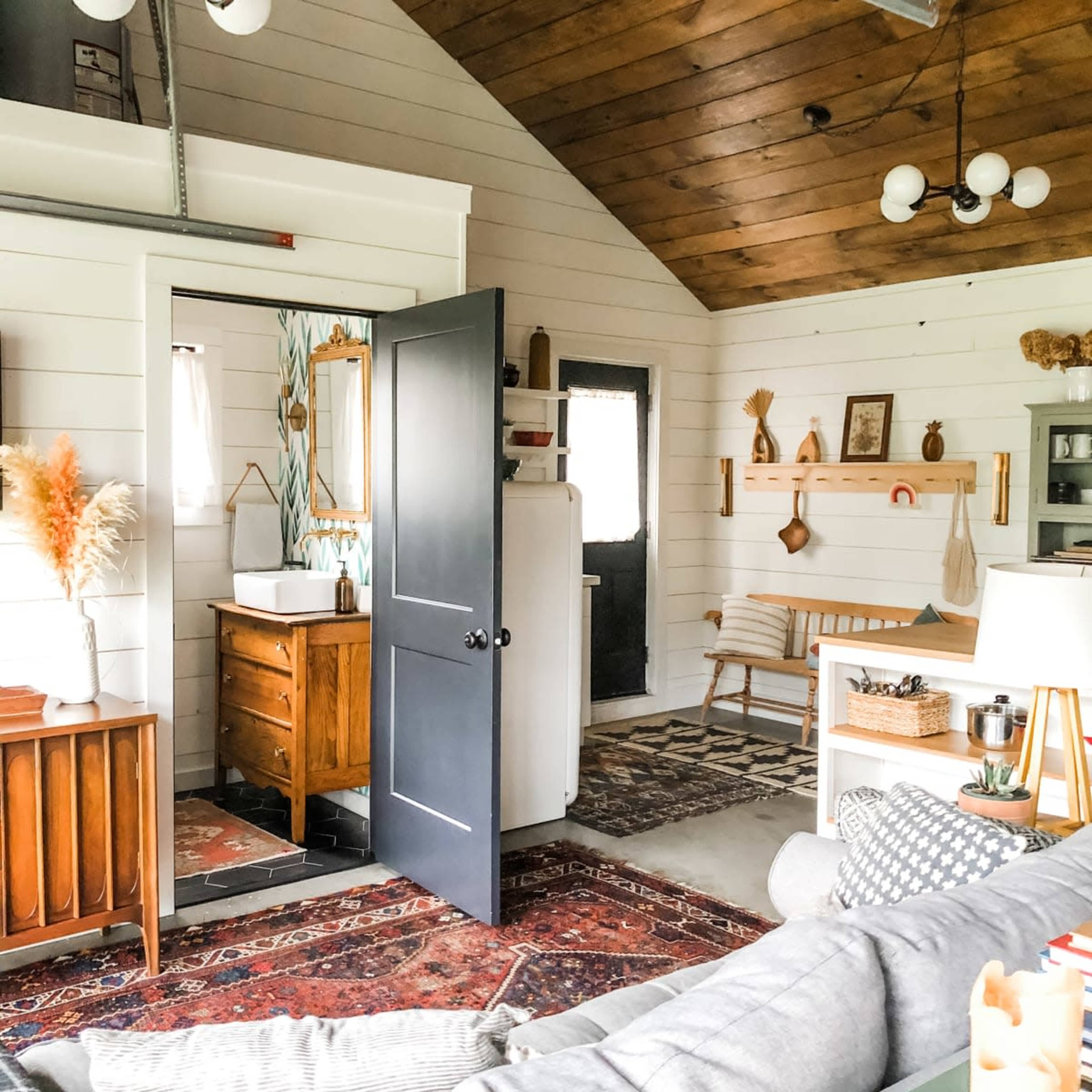The image shows a cozy, well-decorated living space with exposed wooden beams, a bathroom area visible through a black door, and various decorative items arranged on the walls and furniture.